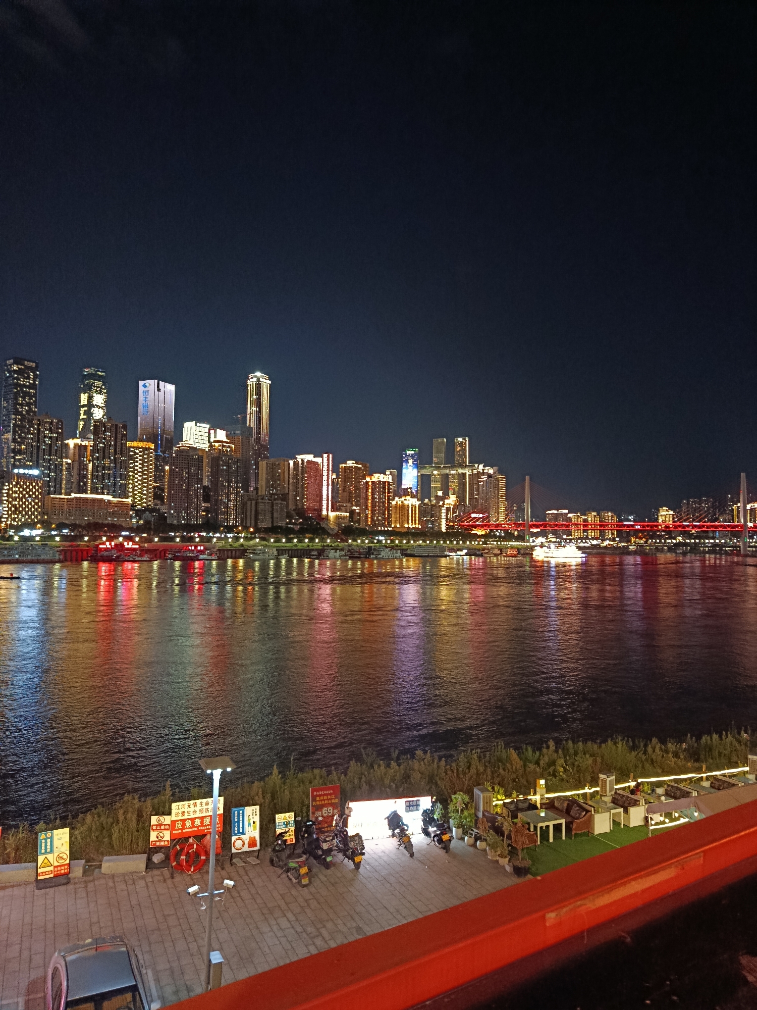 This image captures a nighttime cityscape in Nan'an District, China, overlooking a wide river. A dense cluster of illuminated skyscrapers forms the skyline on the opposite bank, their lights reflecting vibrantly on the water's surface. Several boats, including a large illuminated vessel, are visible on the river. The foreground features a paved walkway along the riverbank, lined with various signs, a life preserver, a lamppost, and several parked motorcycles. To the right, an outdoor seating area with tables and chairs, furnished with plants and decorative lighting, is situated on a raised platform. The overall scene depicts a bustling urban environment at night, with a focus on the illuminated architecture and the tranquil expanse of the river.