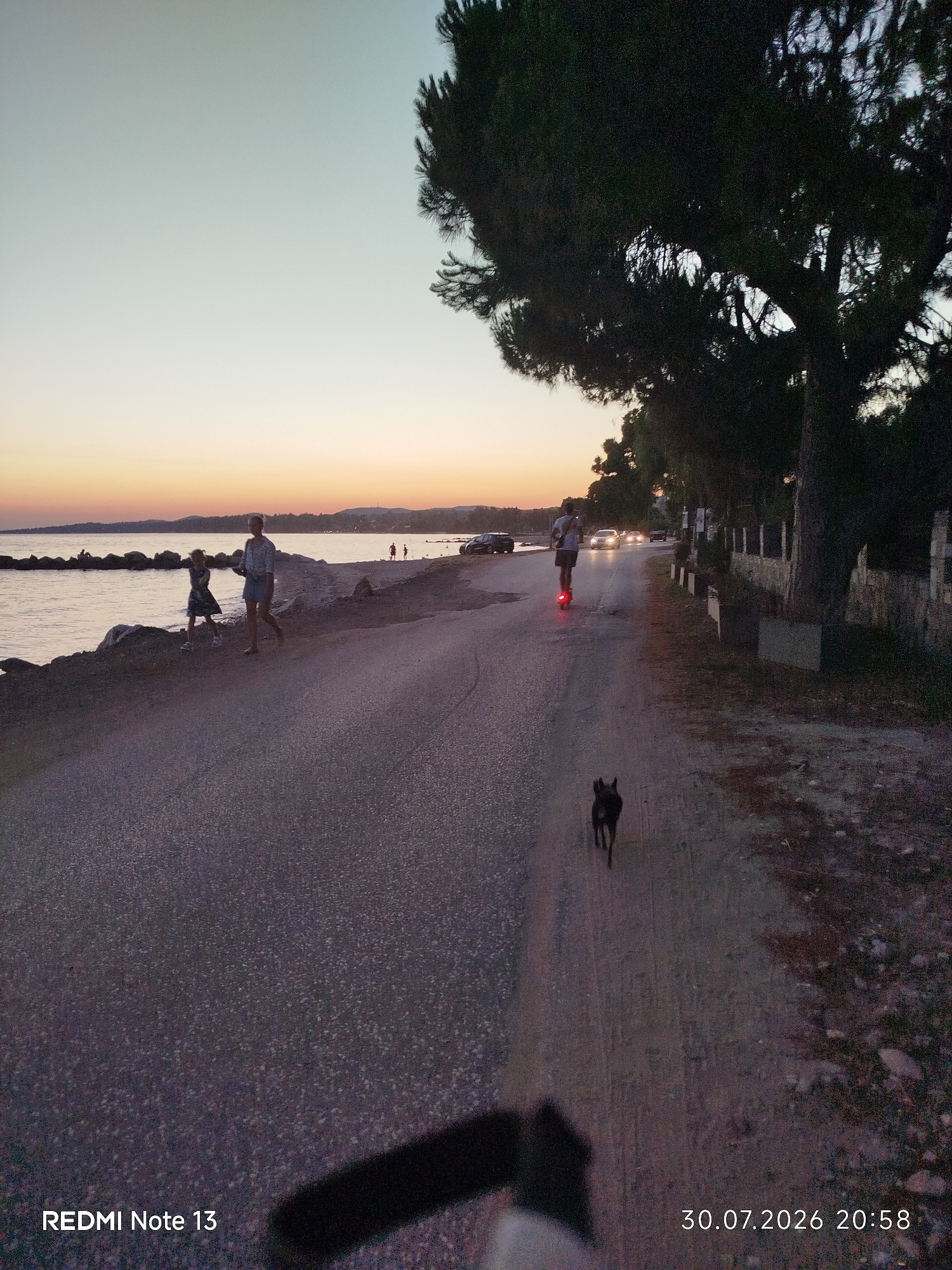 The image depicts a seaside road in Sithonia Municipal Unit, Greece, during sunset. A road with asphalt and gravel runs parallel to a rocky shoreline with calm water. Two young girls, one in a dark dress and the other in a skirt and top, are walking on the gravel shoulder closest to the water. Further down the road, a person is riding an electric scooter, illuminated by its red taillight. A small black dog is running on the road ahead of the scooter rider. Trees, including a large pine tree, line the right side of the road, with a stone wall behind them. Several cars with their headlights on are visible in the distance on the road. The sky exhibits a gradient of pale blue to orange as the sun sets. The bottom left of the image displays "REDMI Note 13" and the bottom right shows "30.07.2026 20:58".