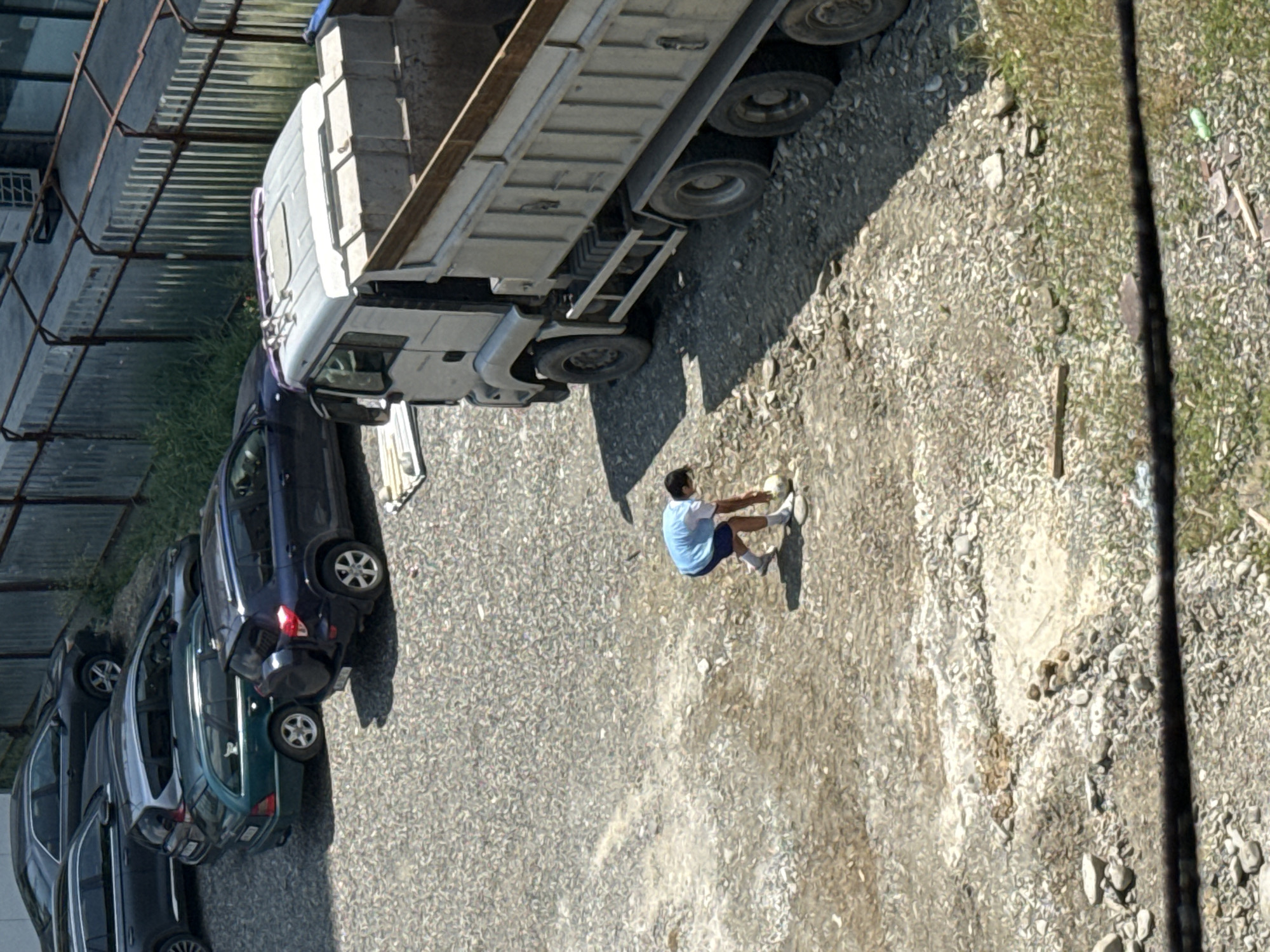 A person in a blue t-shirt and white shorts is bending down to pick up a white ball in a gravel lot. The person is casting a shadow to the left. Behind the person, a white dump truck is parked. Several cars are parked to the left of the truck. A metal fence with green grass behind it runs along the back of the lot. The scene appears to be in Tbilisi, Georgia.