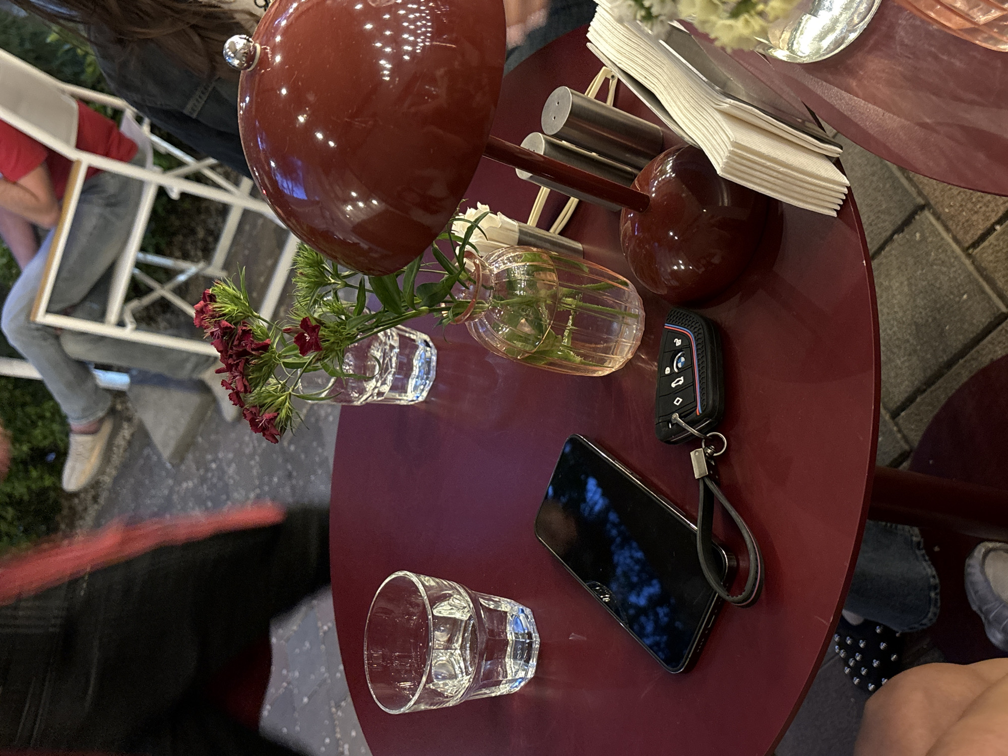 A high-angle shot depicts a maroon-colored round table in an outdoor setting, likely in Yerevan, Armenia. The table holds several items including two small glass vases with flowers, a black smartphone, a car key, a drinking glass with water, a napkin holder, and salt and pepper shakers. Parts of chairs and people are visible around the table, suggesting an outdoor dining or cafe setting. The environment is slightly blurry, focusing on the table's contents.