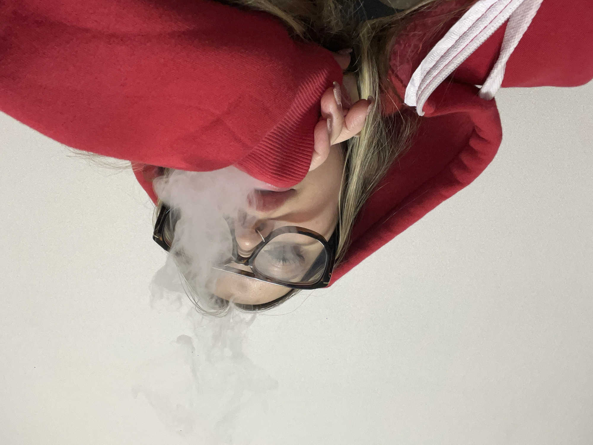 The image, taken in Krasnodar, Russia, captures an inverted, close-up view of a person exhaling a large, dense cloud of white vapor or smoke. The subject, appearing to be female, is positioned against a plain white background and is seen from a low angle. She has long, light-colored hair and is wearing a vibrant red hooded sweatshirt with prominent white drawstrings around the neck and ribbed cuffs on the sleeves.
The person is wearing dark-rimmed glasses, which have a metallic or light-colored bridge and lower frame, resting on her nose. The exhaled vapor billows around her face, partially obscuring her mouth and nose. One hand is visible, with manicured fingernails, positioned near her face and partly covered by the red sleeve. No specific time of day or external environmental factors are discernible from the plain background. There is no visible text within the image. The focus of the scene is primarily on the act of exhalation and the flowing vapor.