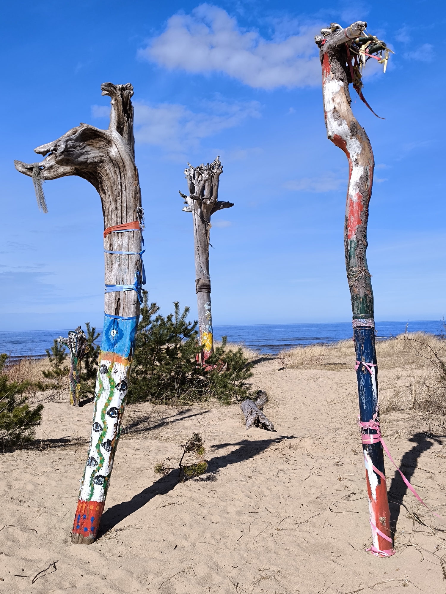 The image depicts a sandy beach with several painted wooden poles or sculptures in Gauja, Latvia. The poles are decorated with different colors and patterns, some with ribbons tied around them. The sky is blue with scattered clouds, and the sea is visible in the background. Pine trees and other vegetation are present among the poles.