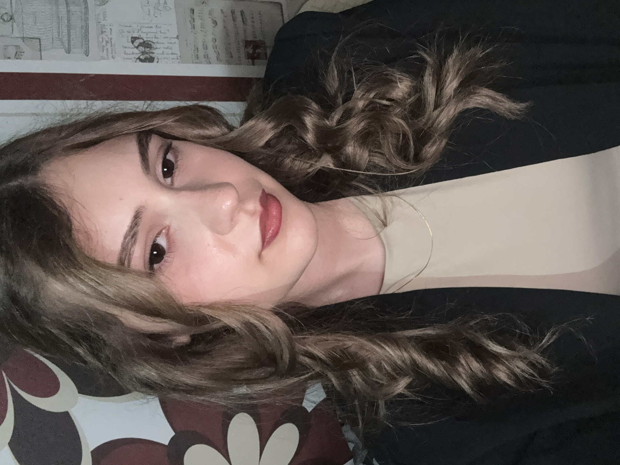 A young woman with long, wavy brown hair is angled towards the camera. She is wearing a beige top under a black blazer. Her makeup is visible, including lipstick. She is positioned in front of a wall decorated with patterned wallpaper. The location in Panagyurishte, Bulgaria, is not discernible from the image.