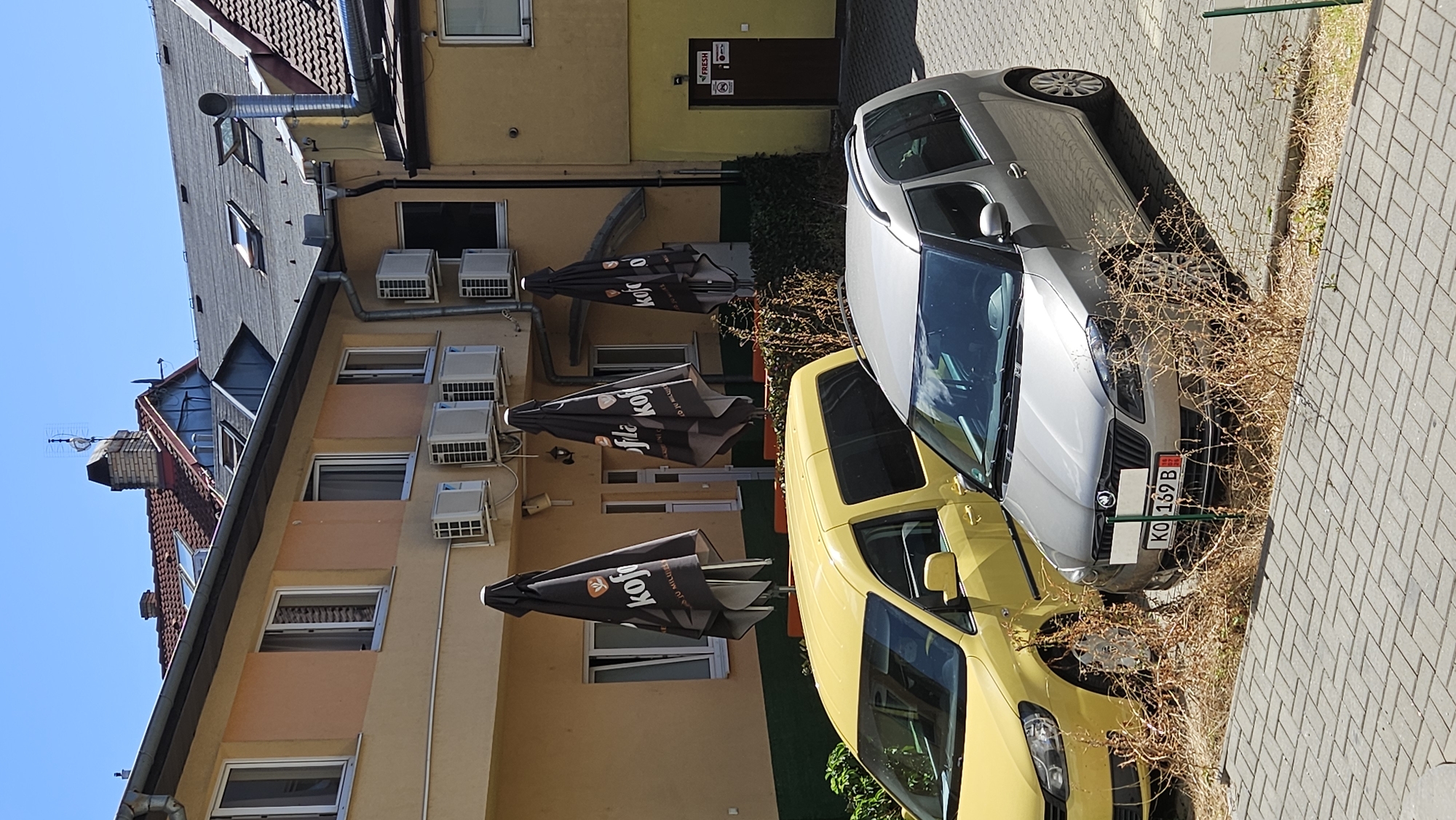 The image shows an outdoor scene in the city of Prešov, Slovakia. In the foreground, two cars are parked: a yellow minivan and a silver hatchback. Both vehicles are surrounded by dry, brown brush. The cars are parked on a paved surface composed of light gray rectangular bricks.
Behind the cars, a two-story building with a yellow facade is visible. Several closed black umbrellas with the logo "kofa" are positioned along the building's exterior. Multiple air conditioning units are mounted on the building's walls. Windows are visible on both floors.
Further in the background, parts of other buildings are visible, including tiled roofs and chimneys. A brown door with a sign that reads "FRESH" is located on the right side of the yellow building. The sky is clear and blue. There are no people visible in the image.