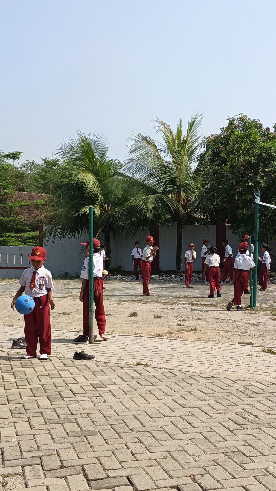 In Pasir Bolang, Indonesia, a group of schoolchildren in red and white uniforms are engaged in an outdoor activity. The scene takes place on a paved courtyard with a dirt area in the background, bordered by a wall and lush green trees, including palm trees. The sky is clear and blue. In the foreground, a young boy wearing a red cap, white shirt with a red tie and emblem, and red pants, holds a blue ball in his left hand. He is standing on the paved area. Behind him, another child in similar attire stands next to a green pole. Several other children, also in uniform, are scattered across the courtyard, some appearing to be playing or observing. A white net is visible in the background, suggesting a sports activity is underway or has just concluded. The overall impression is one of a school sports event or physical education class on a sunny day.