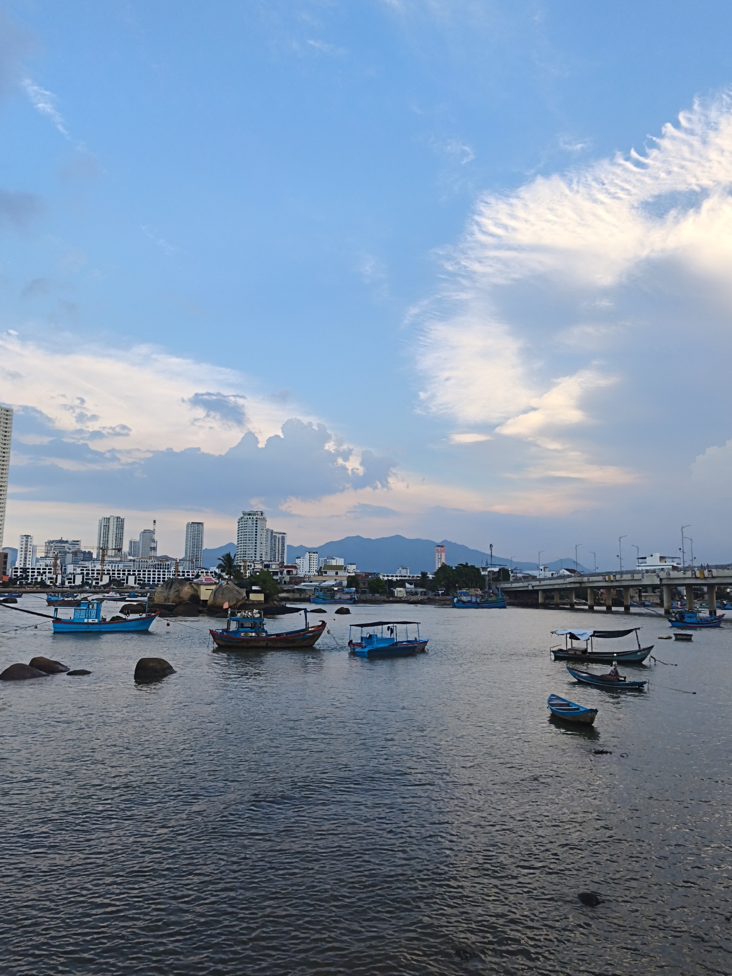 In the city of Bac Nha Trang Ward, Vietnam, a body of water is dotted with several small fishing boats. In the foreground, the water is rippled and dark. In the middle ground, numerous boats, both blue and wooden, are anchored or moving slowly. Some of these boats have canopies. In the background, a cityscape with tall buildings and a bridge can be seen. The sky above is a mix of blue and white clouds, with a hint of pink suggesting it might be near sunset or sunrise. The scene appears to be a harbor or a bay where fishing activities take place.