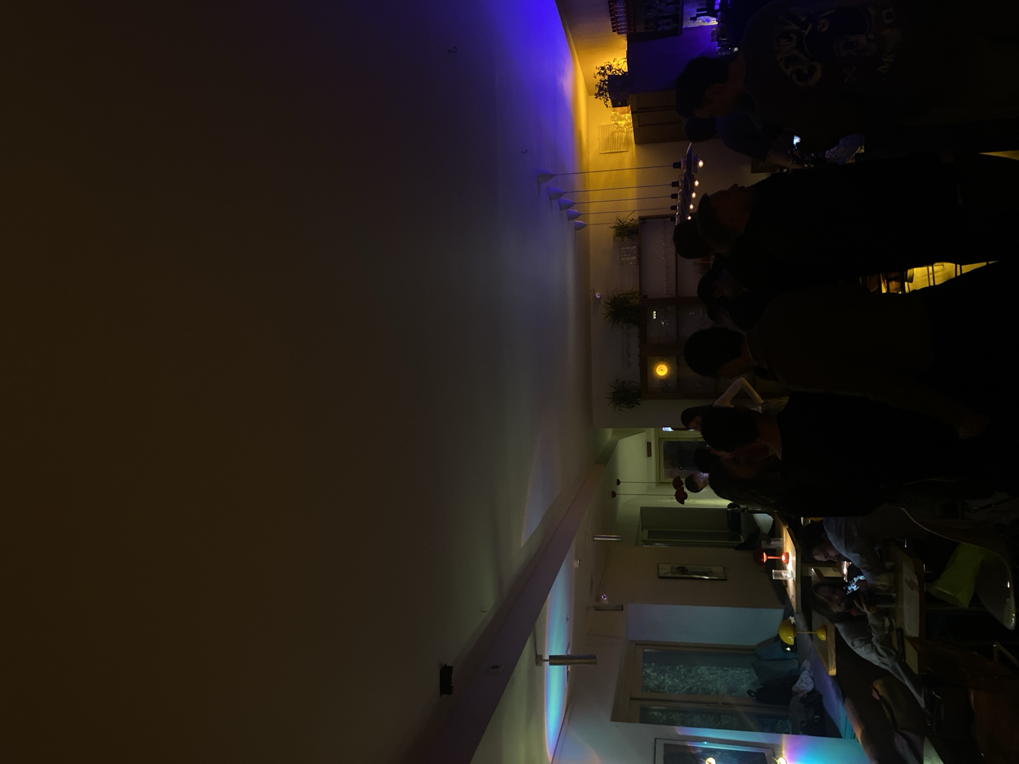 The image shows a restaurant or bar interior. A group of people stands in the right side of the image, at the counter. The interior is bathed in dim light, with yellow and blue hues illuminating the walls and ceiling, and windows are seen in the background. The presence of bottles on shelves near the ceiling suggest a bar area. There are some plants in shelves and along the wall. The location is in Krasnoyarsk, Russia.
