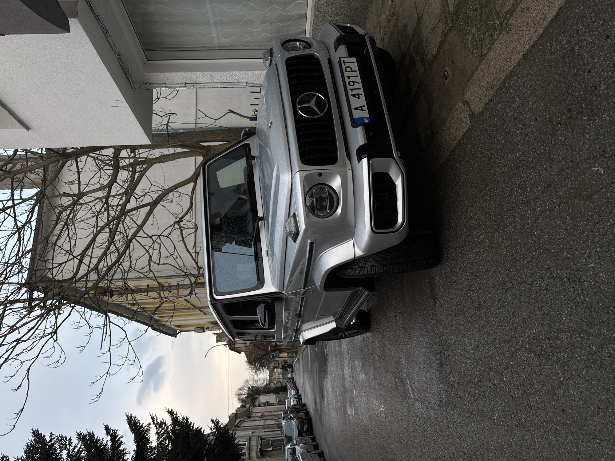 A silver Mercedes-Benz G-Class SUV is parked on the side of a wet asphalt street in Burgas, Bulgaria. The car's license plate reads "A 4191 PT" with "BG" for Bulgaria visible. The vehicle is parked close to the curb, near a building with a white exterior. Bare tree branches are visible in the background against a cloudy sky. Other parked cars line the street further down. The scene is urban, with residential or commercial buildings visible in the distance.