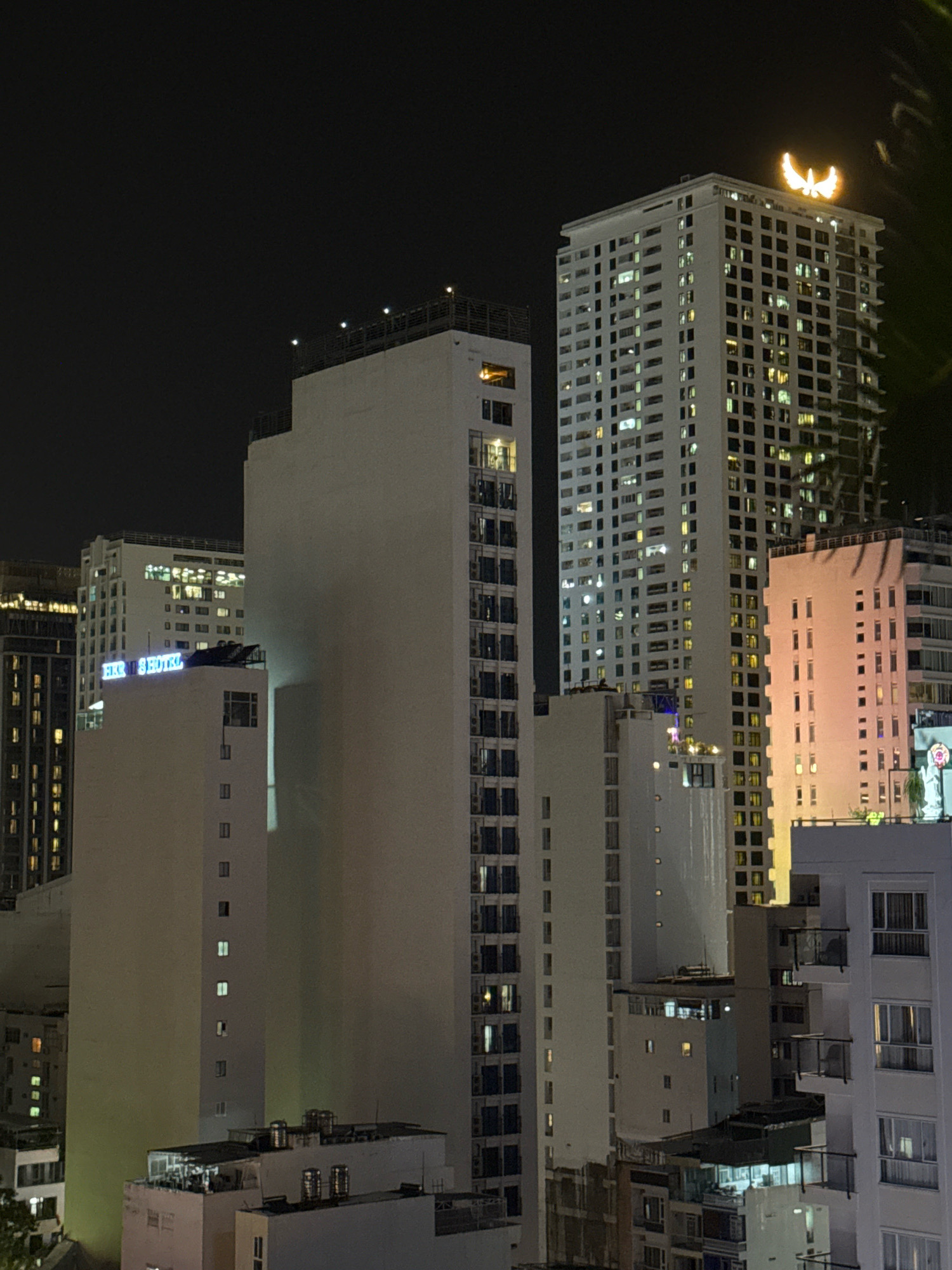 This is a night-time, eye-level, medium shot of a city skyline in Nha Trang, Vietnam. Multiple tall buildings are visible, with lights on in many of the windows. One prominent building in the center features a large, illuminated logo resembling a bird or wings at its apex. To the left of this central building, a smaller building displays blue neon signage that reads "FIKK 135 HOTEL". The buildings are a mix of modern architecture, with some appearing to be hotels or residential high-rises. The sky is dark and clear. There are no people or specific actions depicted in the image, creating a static scene of urban architecture at night.
