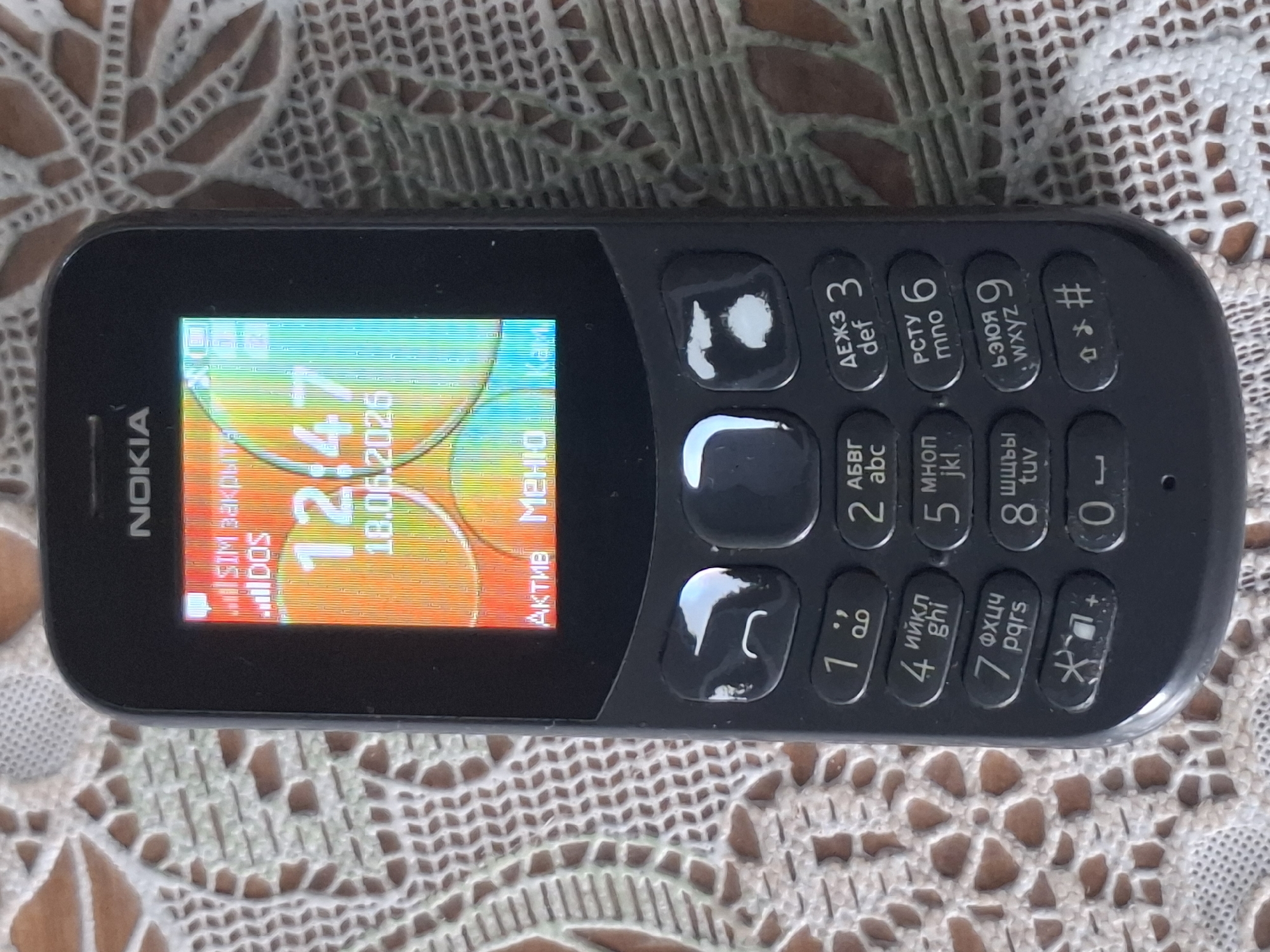 A close-up, eye-level shot shows a black Nokia mobile phone resting on a patterned fabric surface, likely a tablecloth. The phone's screen displays the time as "12:47" and the date "18.06.2026". The screen also shows signal strength icons and text that appears to be in Russian, possibly indicating "SIM" and "Active". Below the screen, the phone's keypad is visible, with numbered keys and other function buttons. The fabric surface has a lace-like, intricate pattern in light brown and beige colors. There is no indication of the city of Almaty, Kazakhstan, or any specific urban environment in this image.