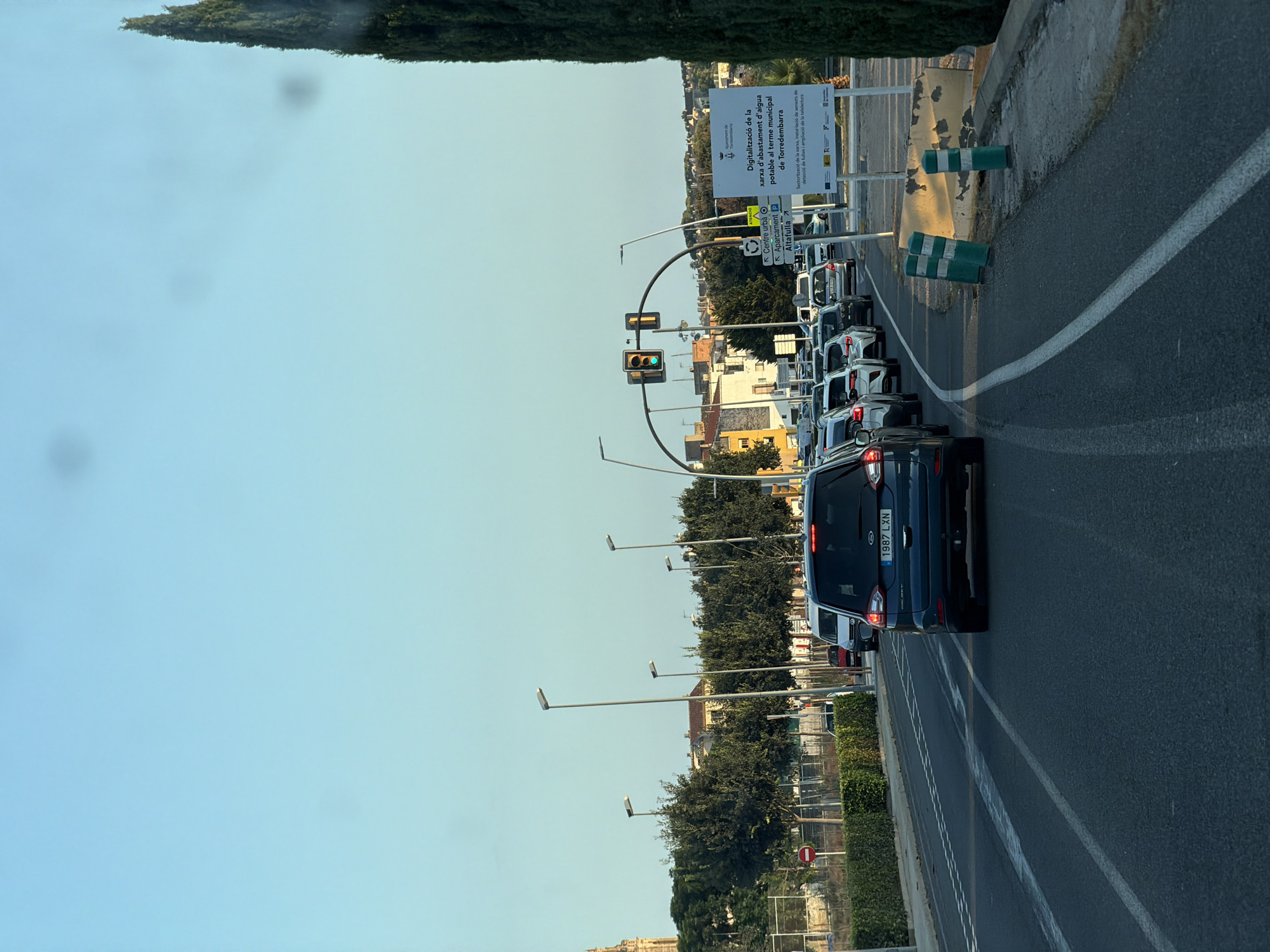 The image depicts a traffic scene on a clear day in Torredembarra, Spain. In the foreground, the rear of a dark blue Ford Galaxy minivan with license plate "1987 LXN" is visible, indicating it is stationary in traffic. Several other vehicles are lined up behind it on a paved road with white lane markings.
To the right of the road, there are several green and white bollards and a concrete barrier. A large white sign with Spanish text and logos is positioned behind the barrier. The text on the sign mentions "Digitalització de la xarxa d'abastament d'aigua potable al terme municipal de Torredembarra" which translates to "Digitalization of the potable water supply network in the municipal area of Torredembarra."
On the left side of the road, a median strip with a low hedge separates the lanes. Beyond the median, there are several streetlights, trees, and buildings. A red circular "no entry" sign is visible on the left. Further in the background, more trees and buildings are present, including what appears to be a church tower with a spire on the far left. The sky is a clear, light blue. The overall scene suggests a traffic jam, possibly at an intersection as indicated by the traffic lights visible in the middle distance.