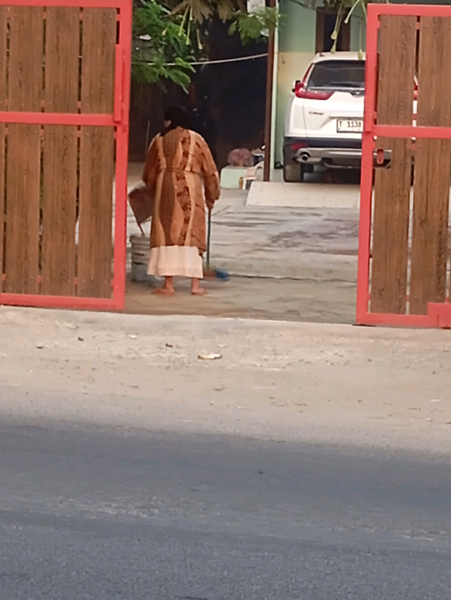 In the city of Babelan, Indonesia, a person, presumably female, is seen from behind, sweeping the ground in front of an open wooden gate. The individual is wearing a long, patterned robe in shades of brown and beige, along with a light-colored skirt and what appear to be bare feet. The person is holding a broom and a dark-colored object, possibly a dustpan or a container, with their left hand. To the right of the person, a white SUV is parked. The scene depicts an everyday activity in an outdoor setting, with a mix of residential and street elements.