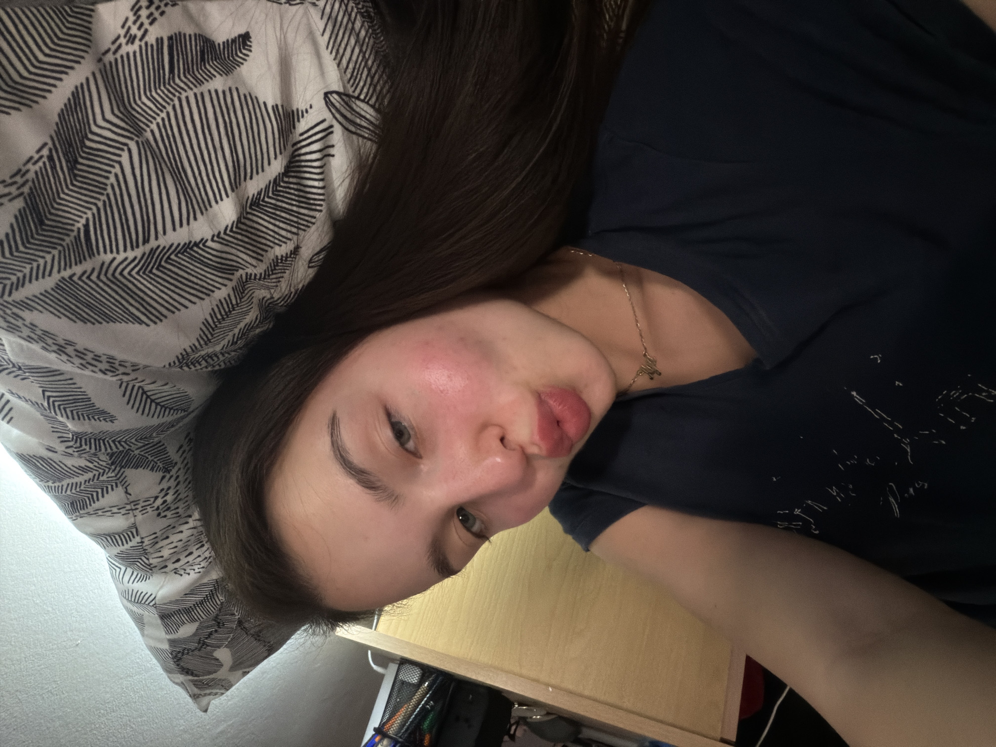 A young woman is lying on her back on a bed with a patterned pillow. She is wearing a dark blue shirt and a gold necklace. She is looking at the camera with her lips pursed. The background is a wall and a desk. The location is Unknown, Unknown.