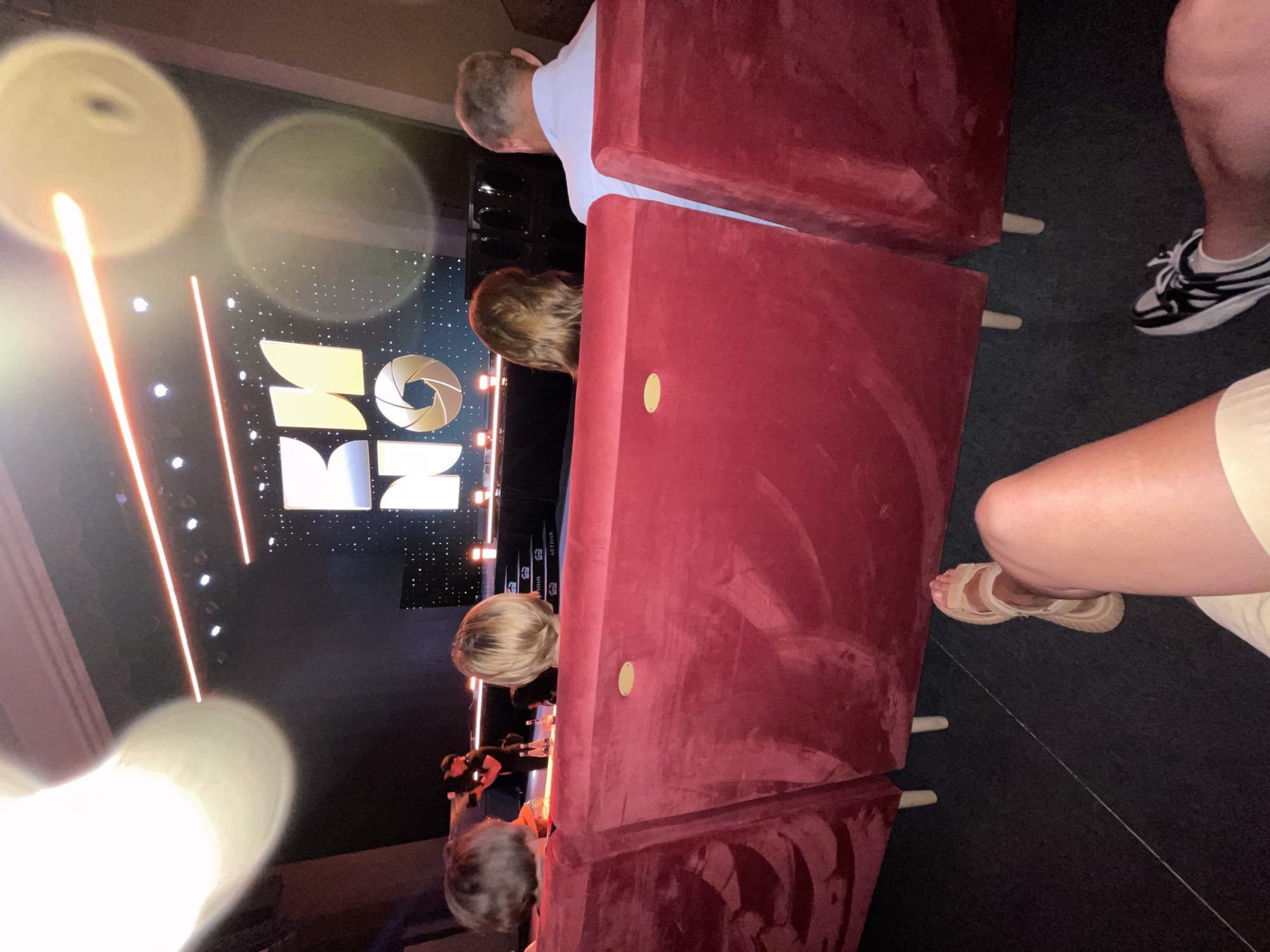 An overhead shot shows a group of people sitting in a movie theater in Sochi, Russia, watching a presentation on a large screen that displays the word "KINO" in large letters. The screen is dark and illuminated by spotlights. The people are seated in red velvet chairs facing the screen, with their backs to the camera. A person in the foreground, on the right side of the image, is wearing sandals and shorts, and another person on the far right is wearing sneakers and pants. The theater appears to be dimly lit, with some light spilling from the screen onto the audience. In the middle ground, on a stage, a person is operating a camera, and there are bottles of water on a table. The overall atmosphere is one of a formal event or screening.