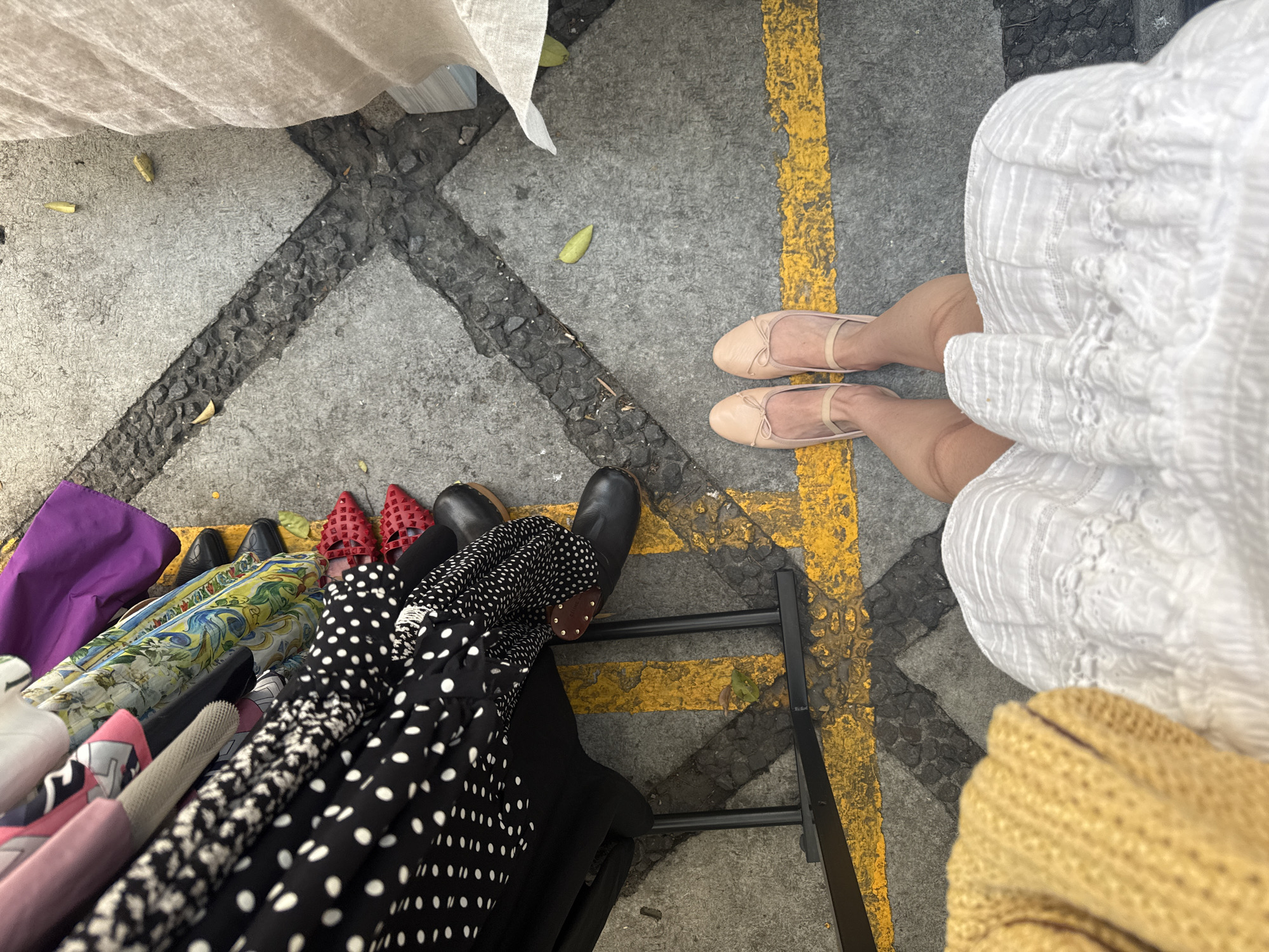 An overhead shot shows the legs and feet of two individuals standing on a concrete surface marked with yellow lines and dark gray square patterns. The individual on the right wears a white dress and light peach-colored ballet flats. To the left, a rack displays a collection of clothing items, including dresses with various patterns (floral, polka dots, black, etc.) and other fabrics in different colors (purple, yellow, etc.). The legs of the other individual are partially obscured by the clothing rack. This scene suggests an outdoor market or street vendor setting. It is likely located in Mexico City, Mexico.