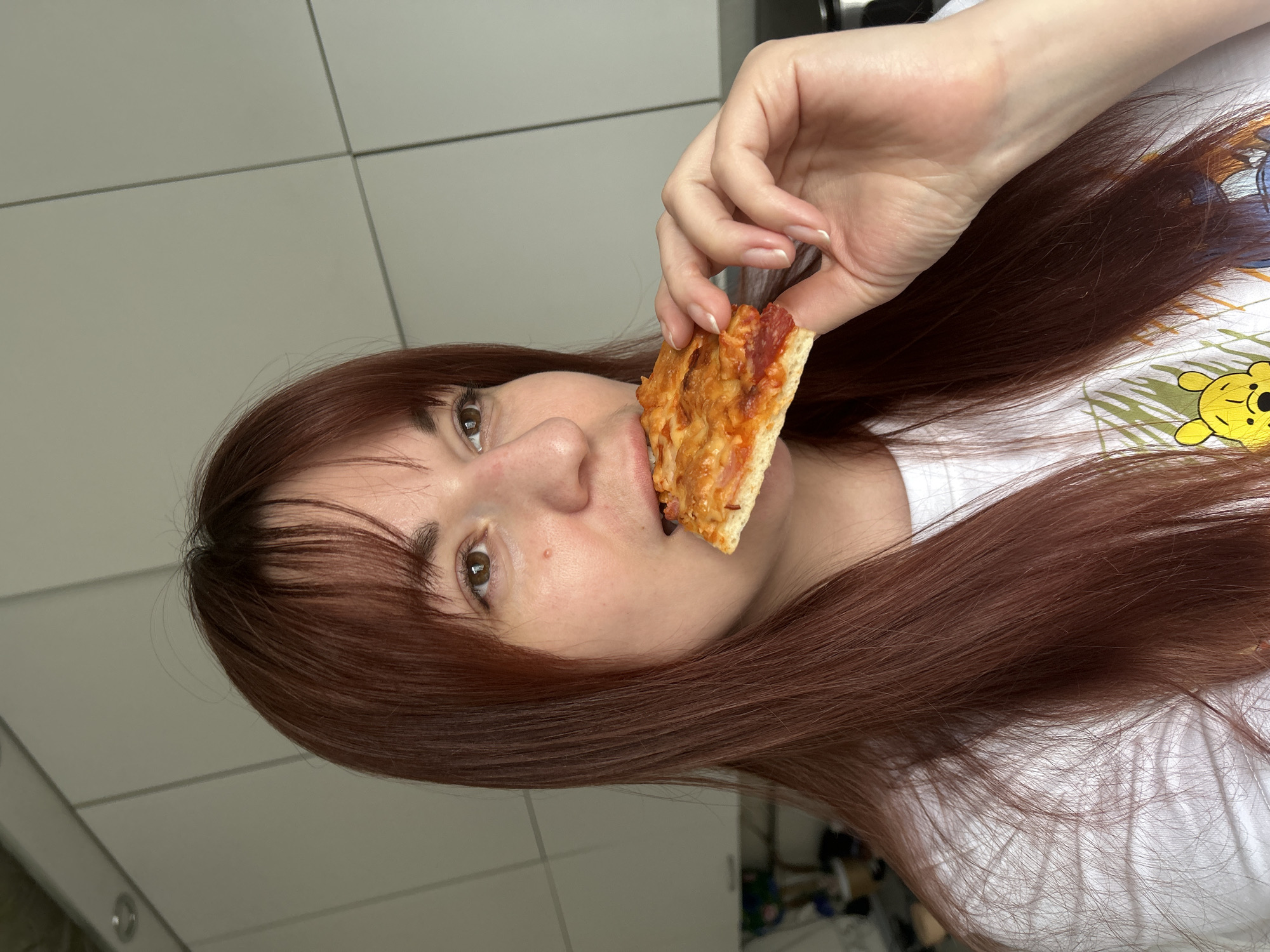 A fair-skinned woman with auburn hair is holding a slice of pizza to her mouth. She is wearing a white t-shirt. The background consists of white tiled walls and an area with indiscernible objects. The location is specified as Фоки, Russia, but there is no visual confirmation of the city in the image.