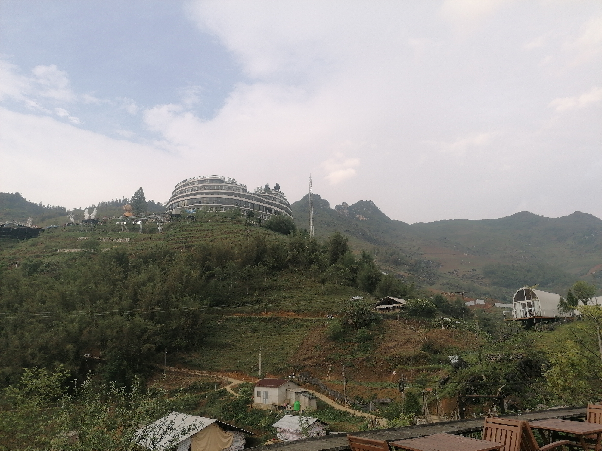 This wide shot shows a mountain landscape in Sa Pa Ward, Vietnam under an overcast sky. A large, multi-story building with a curved facade sits atop a green hill. In the background, there are mountains, one with a tall telecommunications tower near the peak. Smaller buildings and structures are scattered on the lower slopes of the hills, including a white dome-shaped building. In the foreground, there are wooden tables and chairs.