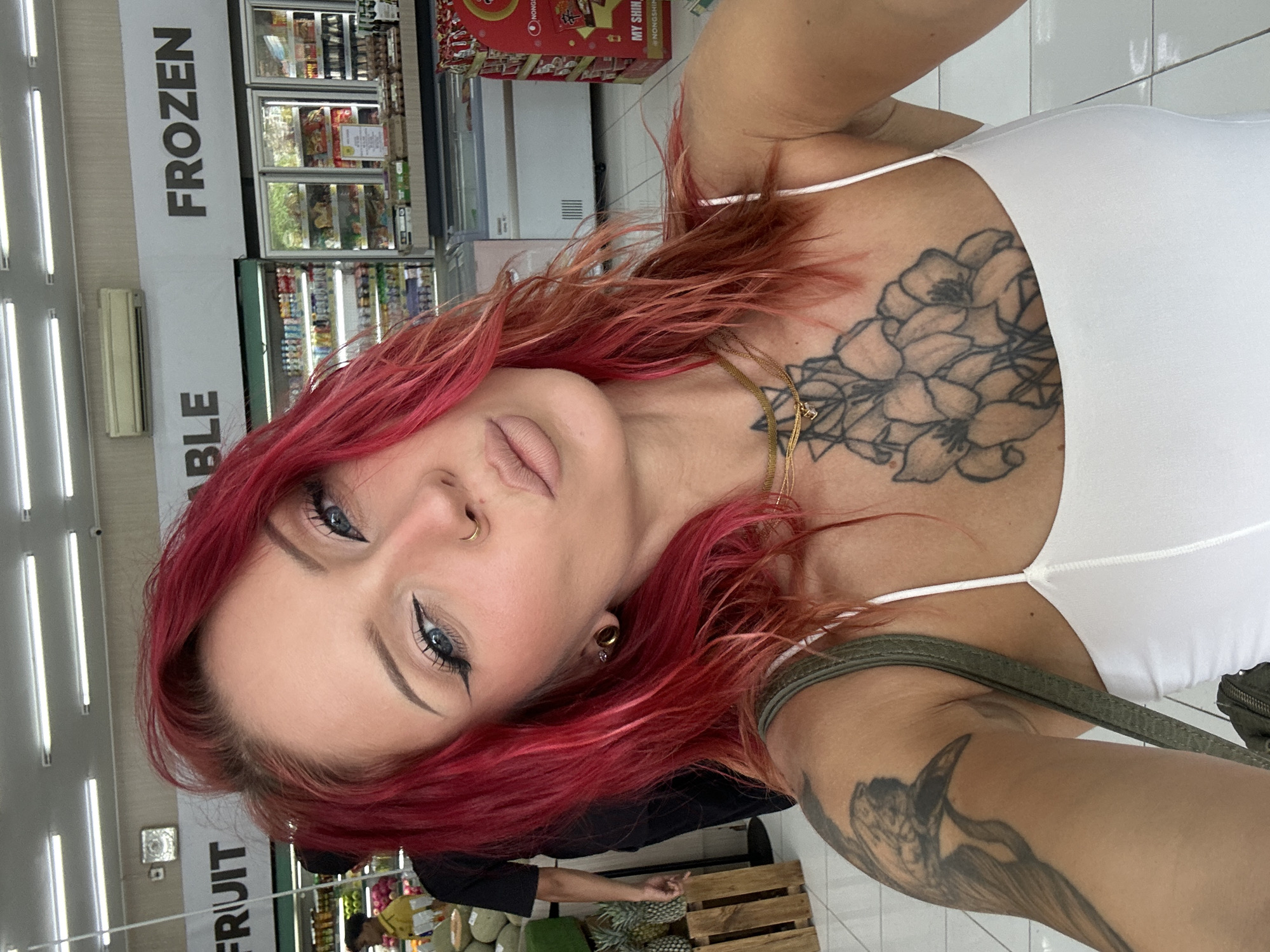 A woman is taking a selfie inside a market. She has red hair and light eyes, and is wearing a white tank top. She has multiple tattoos. The market has signs for "FROZEN" and "FRUIT" in English. The location is Kuta Selatan, Indonesia. Pineapples are visible.