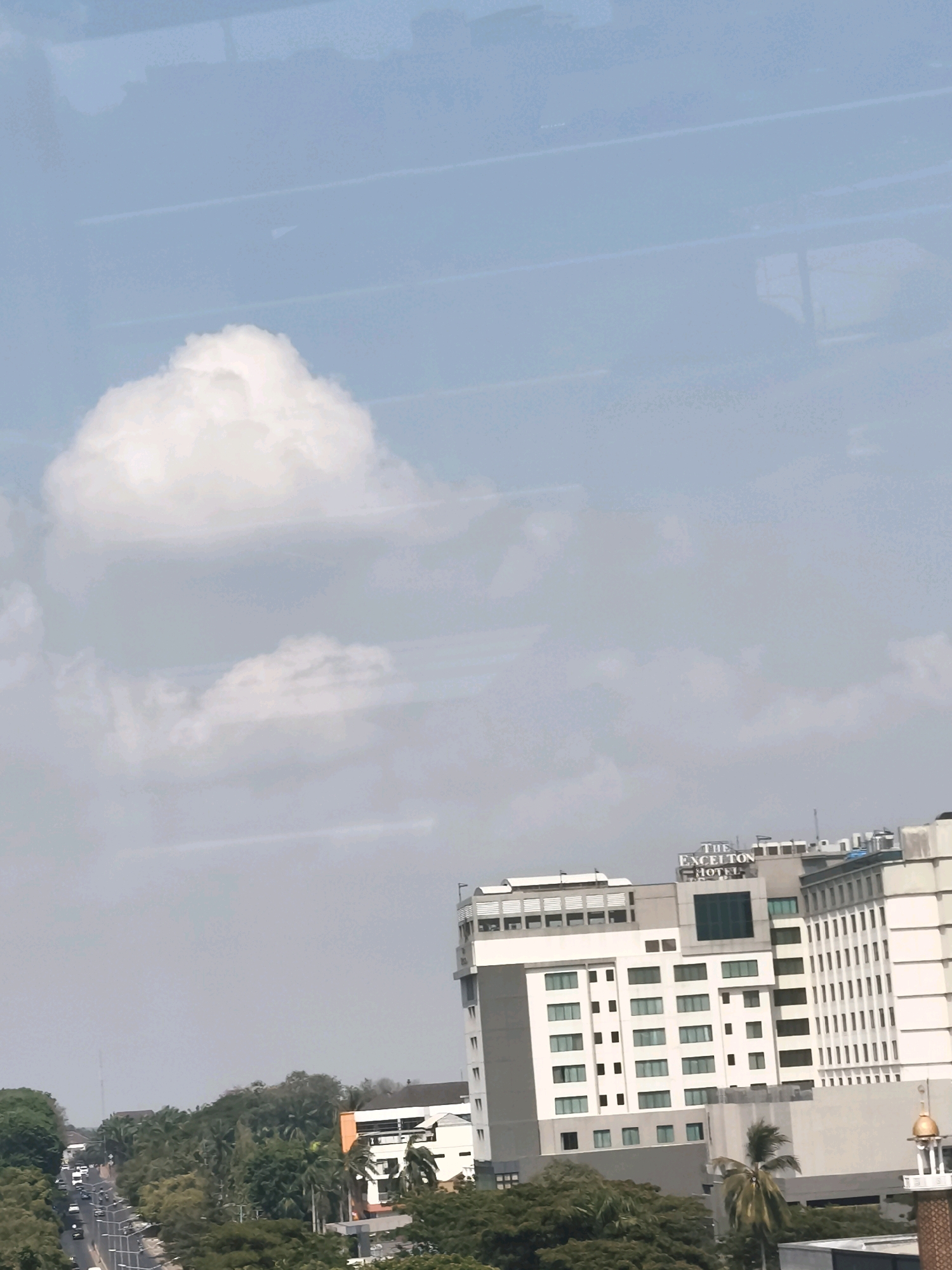 The image depicts a daytime aerial view of a city, likely Palembang, Indonesia, focusing on a street scene with buildings and vegetation. In the foreground, a large, modern hotel complex, identified as "THE EXCELTON HOTEL" by signage, dominates the right side of the frame. The hotel is a multi-story structure with a white facade and numerous windows. To the left of the hotel, a tree-lined street can be seen with several vehicles in motion, suggesting ongoing traffic. The middle ground is characterized by dense greenery, comprising various trees and foliage. In the background, the sky is partly cloudy, with large, fluffy white clouds against a light blue backdrop. The overall impression is one of a bustling urban environment with significant green spaces.