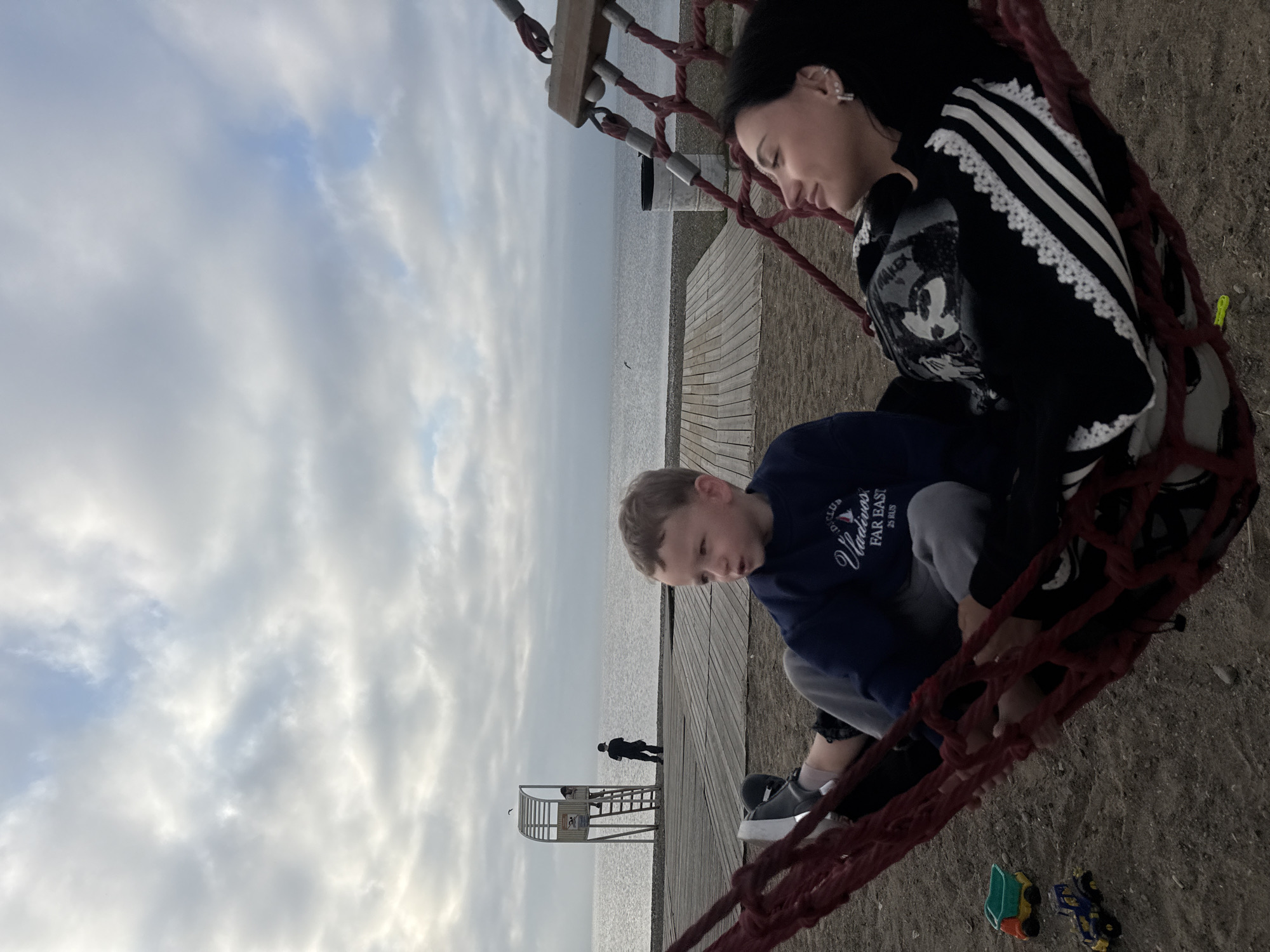 A young boy and a young woman are relaxing in a red net hammock on a sandy beach overlooking the sea. The boy is wearing a dark blue sweatshirt with "Vladivos FAR EAST 25 RUS" printed on it in white. He is looking to his right, with a slight smile. The woman is lying next to him, wearing a black jacket with white stripes and lace trim. She is looking up and to her right, with a gentle smile. In the background, there is a wooden pier and a lifeguard tower. The sea is calm, with a cloudy sky overhead. Several toy vehicles, including a green and orange truck and a blue and yellow tractor, are scattered on the sand near the hammock. The location is the beach in Vladivostok, Russia.
