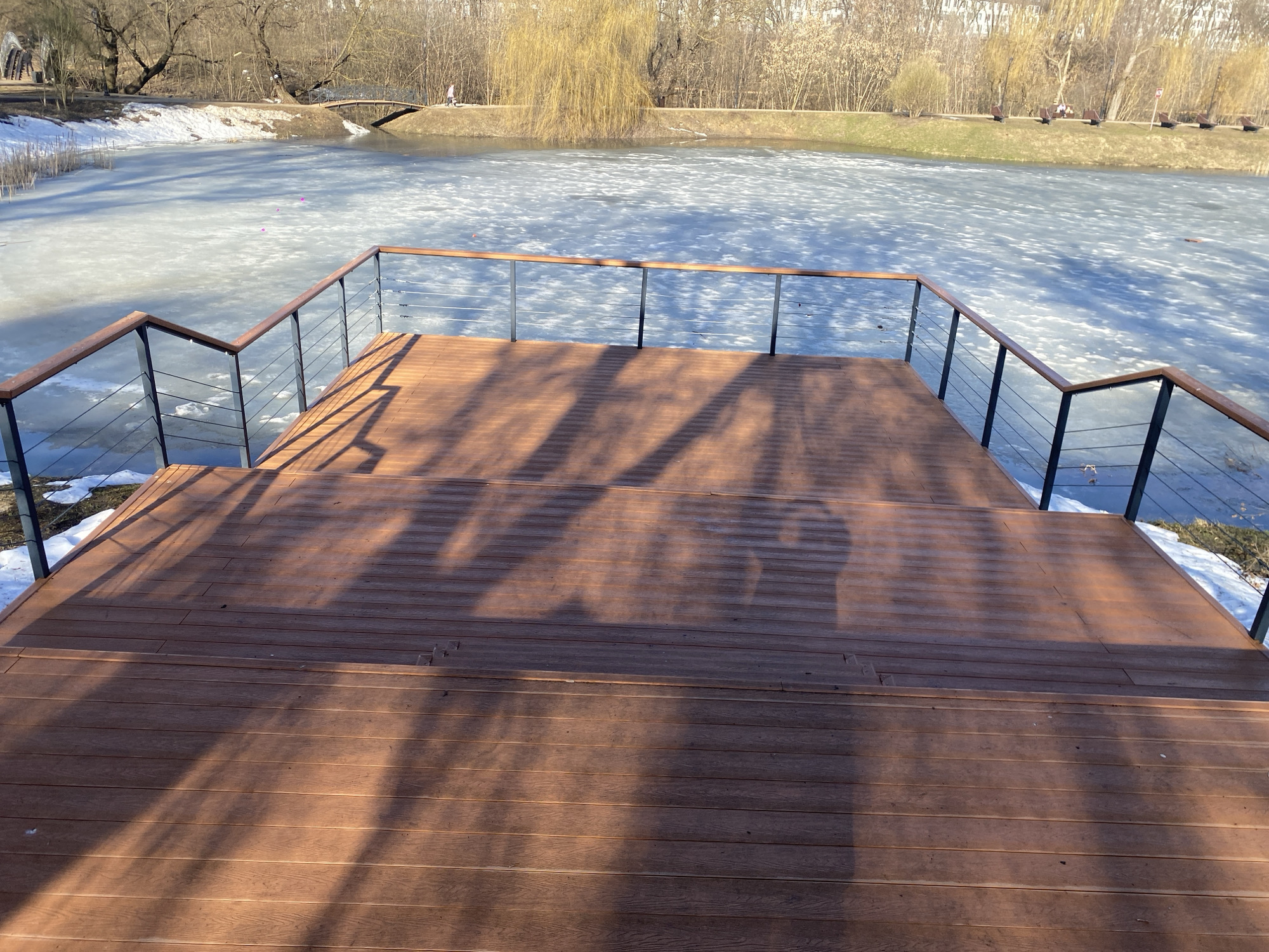 The image captures a scene at the Tsaritsyno Park in Moscow, Russia, during what appears to be late winter or early spring. A wooden deck with metal railings overlooks a partially frozen lake. Remnants of snow are visible on the banks. The ice on the lake is thawing. A small arched bridge is seen in the background. Trees without leaves, along with some evergreens, surround the lake. The scene is captured in daylight, with shadows cast on the wooden deck. The shadows appear to be those of people.