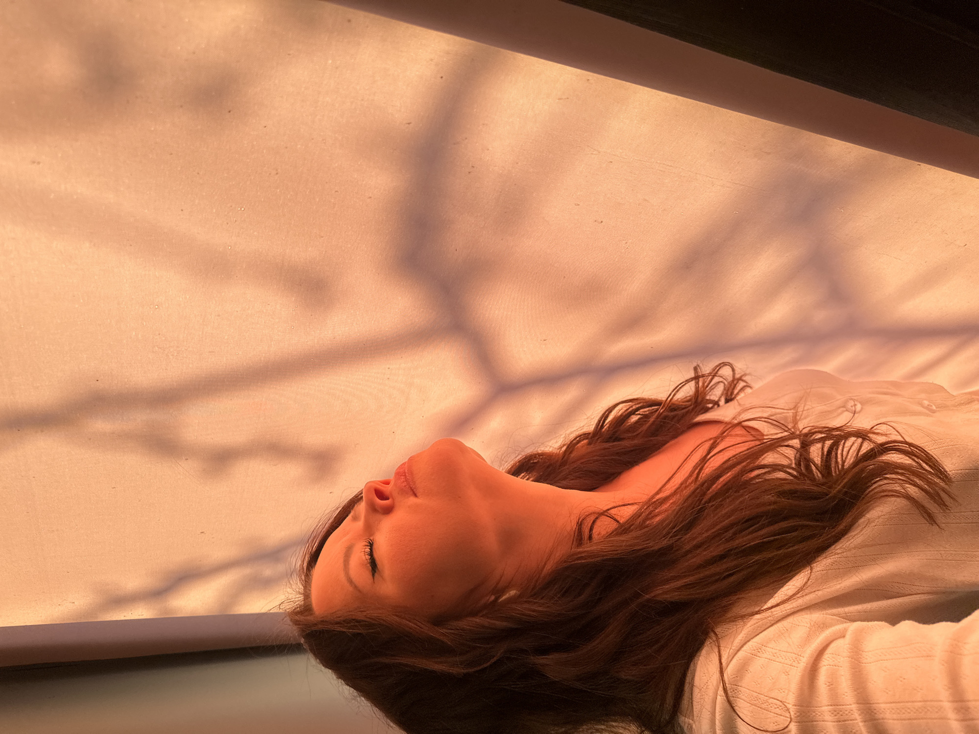 A low-angle shot depicts a woman lying on her back, her head tilted upward towards a sunlit fabric-covered surface. The light casts shadows resembling tree branches onto the fabric. She has long, dark brown hair that spreads across her white top. The focus is on her face, particularly her eyes, nose, and mouth. The scene appears to be indoors with light filtering through a translucent material. The location of Chaikovsky, Russia, cannot be independently verified using image data alone.