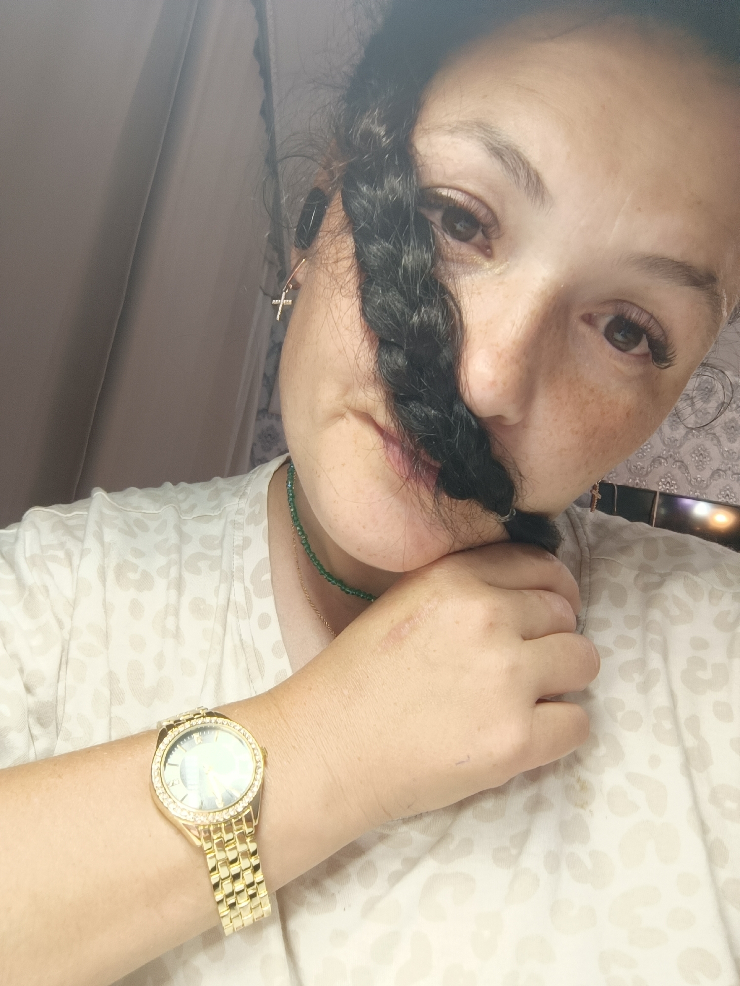 The image is a close-up, head and shoulders selfie of a woman. She is wearing a light-colored t-shirt with a subtle leopard print pattern. On her left wrist, she has a gold-colored watch with a diamond-encrusted bezel and a light-colored face. She is also wearing a green beaded necklace. A dark braid of hair is draped across the front of her face, partially obscuring her features. A small cross earring is visible on her right ear. The background is out of focus and appears to be indoors, with a blurred curtain or fabric on the left and patterned wallpaper or decor on the right, along with what looks like a lit object. The lighting suggests it is daytime or a well-lit indoor space. There are no indications in the image that this scene is located in the city of Chaikovsky, Russia.