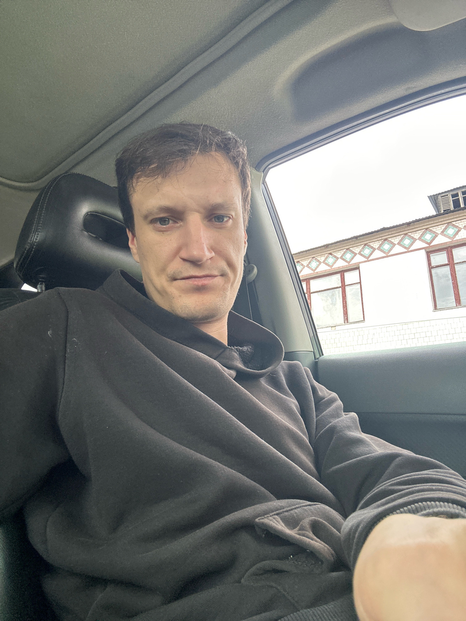 A medium shot shows a man in a black hoodie sitting in the passenger seat of a car, facing the camera. He has short, brown hair and is looking directly at the viewer with a neutral expression. The interior of the car is visible, including the headrest and a portion of the sunroof. Through the car window, a building with a light-colored facade and patterned trim is visible, with a few windows. The sky appears overcast. The man's right arm is resting on his lap. The scene is likely in an outdoor urban or suburban setting.
