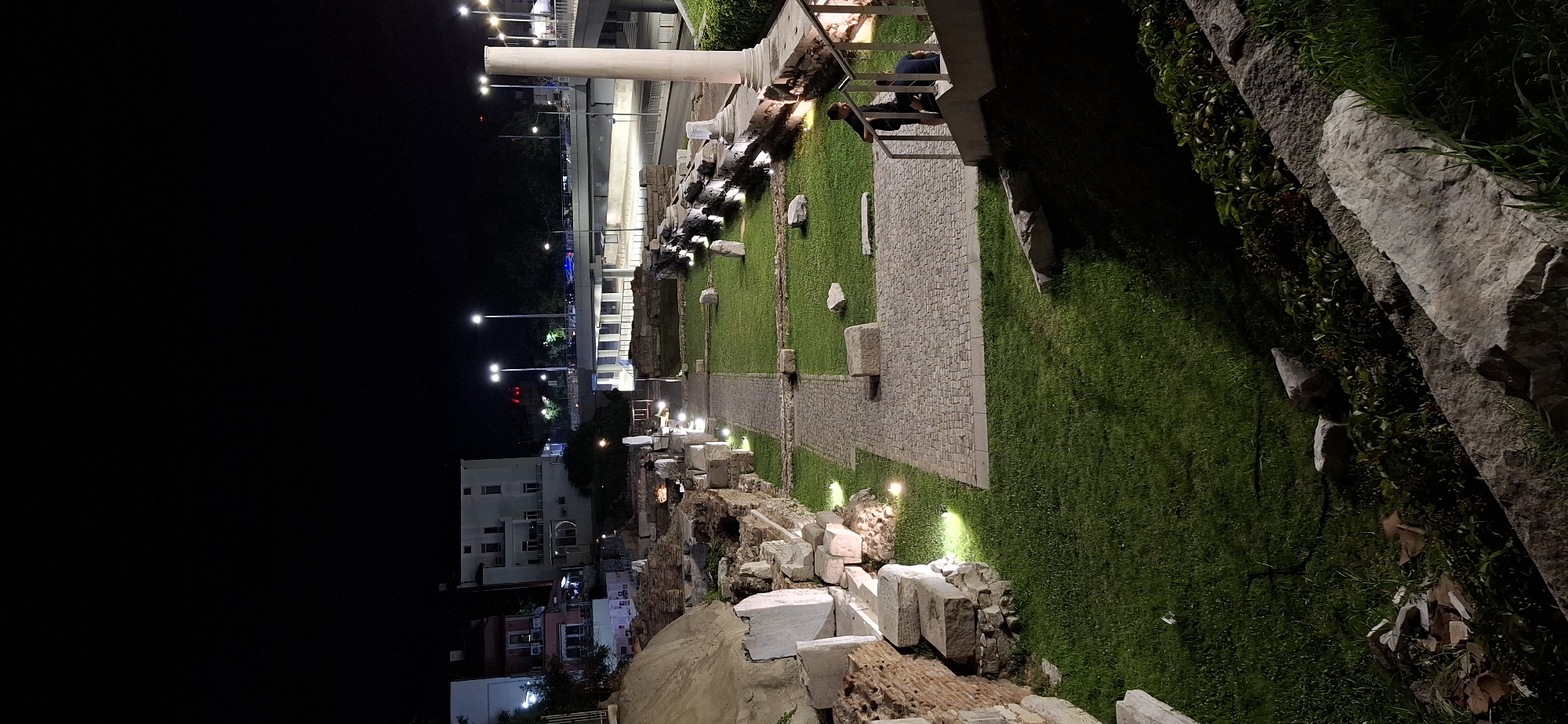 The image depicts an illuminated archaeological site at night in Plovdiv, Bulgaria. The site features ancient ruins composed of stone blocks and brickwork, overgrown with grass in some areas. Paved pathways wind through the ruins, and several large stones are scattered across the lawn. A tall, fluted column stands prominently on the right side of the image, partially illuminated. In the background, modern buildings and an elevated road with traffic are visible. Two individuals are standing near a staircase on the right, partially obscured by shadows. The scene is lit by artificial lights strategically placed around the archaeological area, creating a dramatic ambiance against the dark night sky.