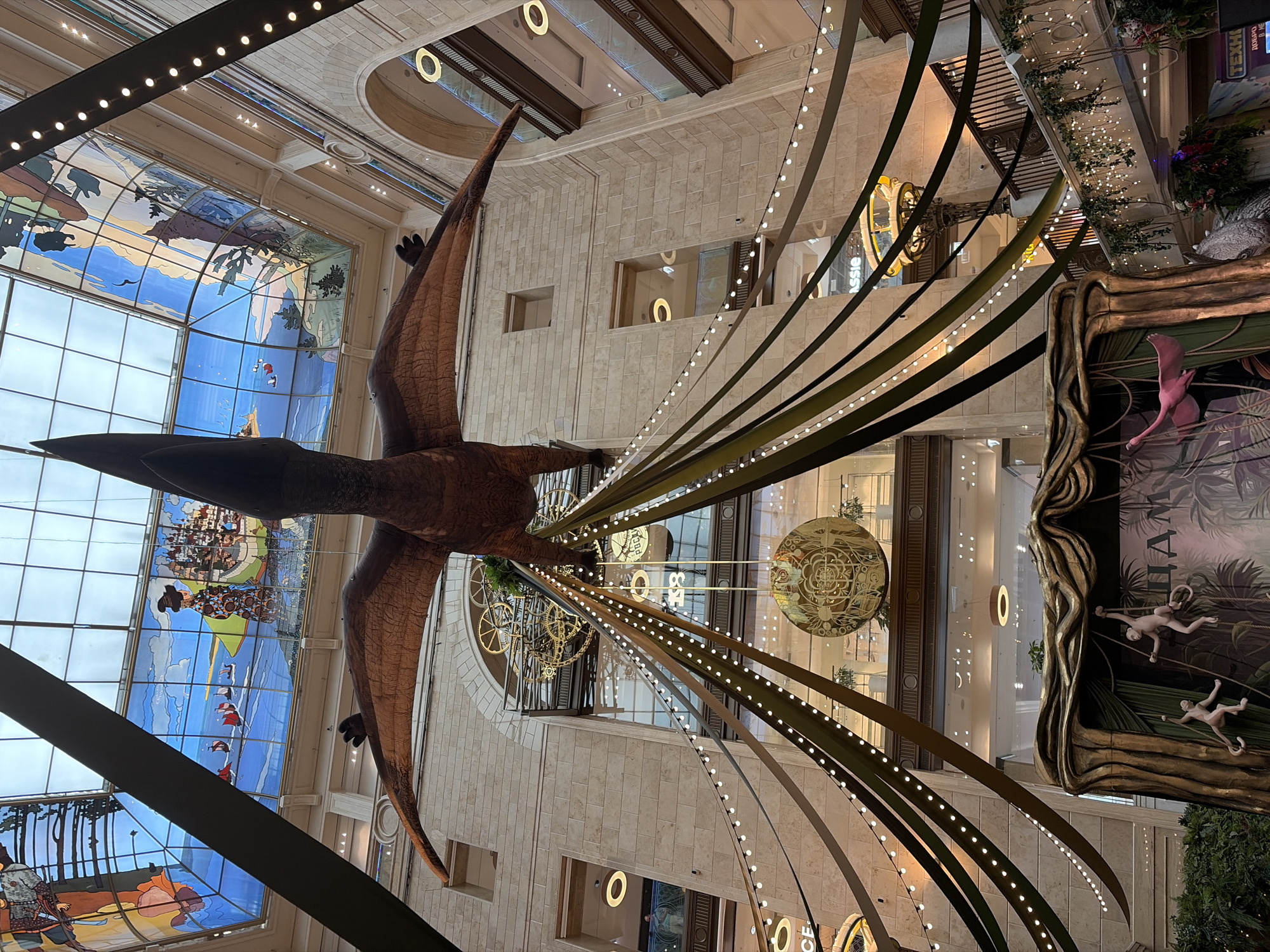 A low-angle, full-shot photograph captures a large, brown pterodactyl model suspended from the ceiling of a grand, multi-level interior space. The dinosaur's wings are outstretched, and its head is tilted upwards. Strands of green and black fabric, interspersed with strings of small white lights, hang down from the pterodactyl's body, creating a festive, almost theatrical atmosphere.
Below the pterodactyl, a circular, golden ornament with intricate patterns hangs, adding to the decorative elements. Further down, a jungle-themed display features several light-colored monkey figures climbing on ropes and vines. A pink flamingo figure is also visible, seemingly in mid-flight. In the background, multiple levels of balconies and windows overlook the central space, with ornate architectural details, including large arched windows and a prominent mechanical clockwork mechanism visible on one of the upper floors. The ceiling is a large, stained-glass skylight depicting a blue sky with clouds, sailing ships, and figures, adding a touch of artistic flair.
The lighting appears to be a mix of ambient interior light and the decorative string lights, casting a warm glow on the scene. The overall impression is one of a large, public indoor space, possibly a shopping mall or an exhibition hall, decorated for a special event or exhibition with a prehistoric and jungle theme. The architectural style suggests a historical building, possibly in a European city. The text "ASSIA" is visible on signage in the mid-ground. The text "ТЕХНО" is visible on signage in the lower right corner.
This scene is located in the historic TSUM department store in Moscow, Russia, which is known for its elaborate seasonal decorations and impressive interior architecture, including its famous stained-glass ceiling.