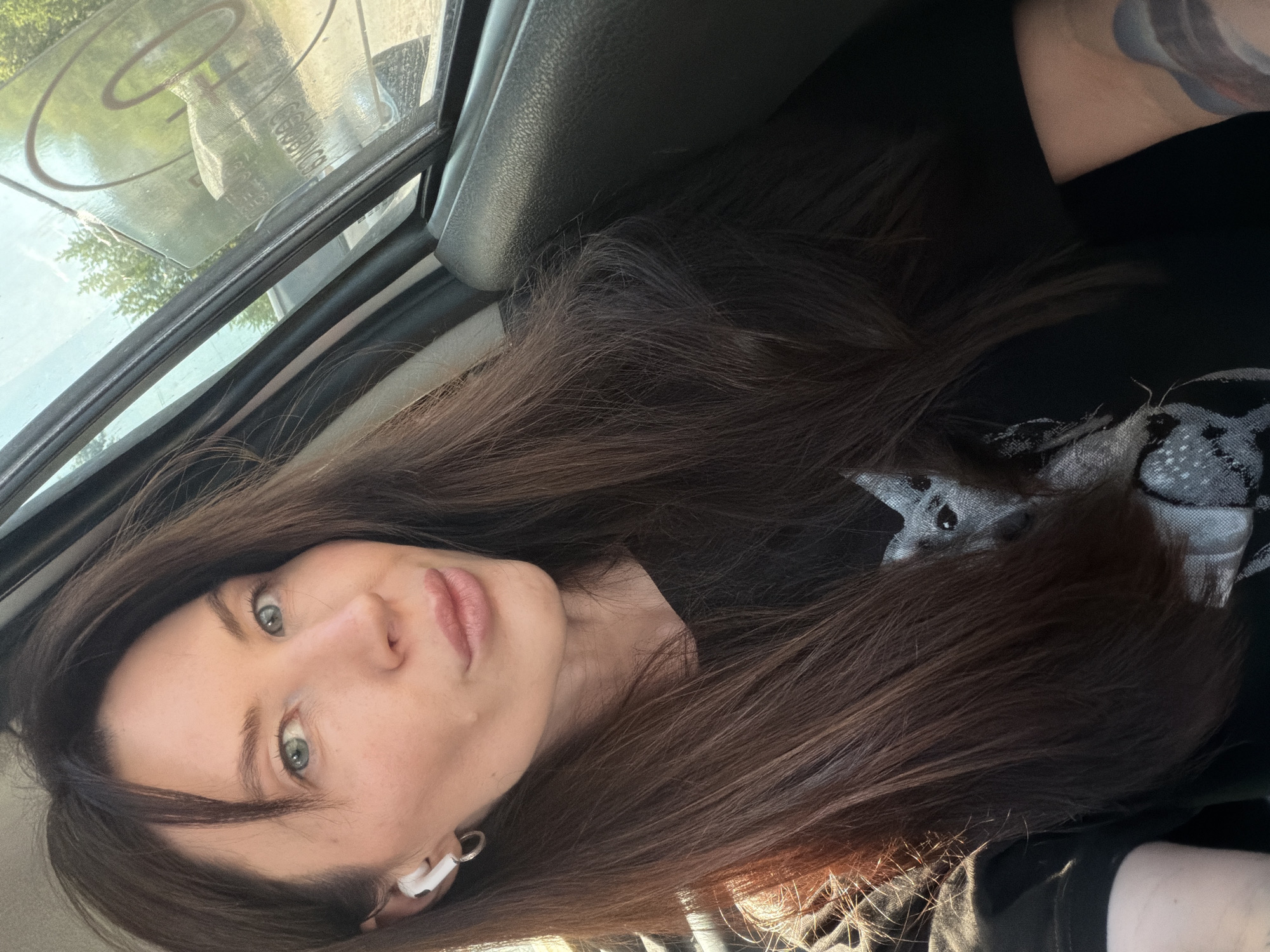A woman with long brown hair and green eyes is inside a vehicle, possibly a car. She is wearing a black t-shirt and has a white earbud in her left ear. The vehicle window shows a blurred view of trees and other objects outside. The location is potentially in Perm, Russia, based on prior knowledge but there are no visible landmarks to confirm that in this image.