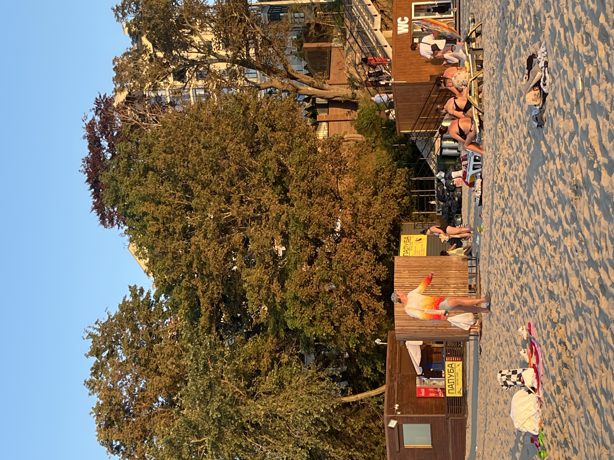 The image depicts a sandy beach in Pionersky, Russia, under a clear blue sky during what appears to be late afternoon or early evening, given the warm, golden light. In the foreground, several beachgoers are relaxing on loungers and the sand. Among them, a person in a multi-colored shirt and gray shorts walks towards the right, carrying bags. Beach towels and personal belongings are scattered on the sand.
Behind the beachgoers, there are wooden structures that appear to be concessions or changing rooms. One structure on the left has a sign with Cyrillic text that reads "ПАЛУБА" (Paluba), suggesting it might be a bar or cafe. Another wooden structure on the right has a prominent "WC" sign, indicating restrooms. A staircase leads up from the beach area towards buildings visible in the background.
The background is dominated by lush green trees, some of which are tinged with autumnal colors, and a multi-story building, likely an apartment complex or hotel. The overall scene captures a leisurely atmosphere on a public beach.