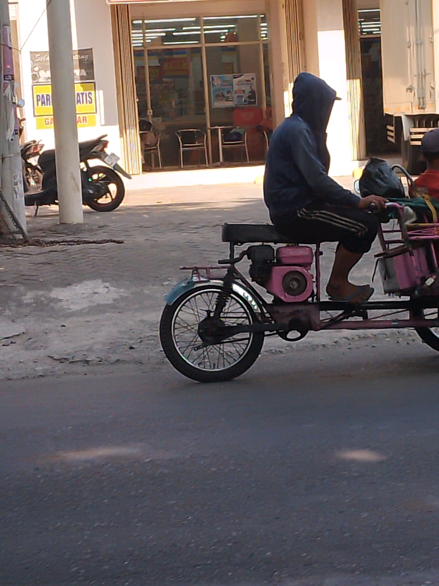 In Kertosono, Indonesia, a person wearing a dark blue hooded jacket and black pants with white stripes is riding a pink and black three-wheeled motorized vehicle. The vehicle has a small engine mounted to its frame. The rider's face is obscured by the hood. The vehicle is on a paved road, with a motorcycle parked to the left and a building with shops visible in the background. The building's facade features signage, including yellow signs with text in Indonesian. The environment appears to be an urban street scene during daylight.