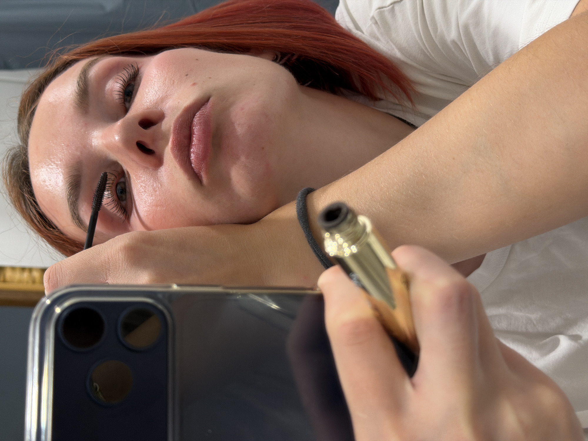 A woman with reddish-brown hair is applying mascara while holding a smartphone. The phone's camera lenses are visible. She is lying down, possibly in an indoor setting. The background is blurred and indistinct. The image's location is unspecified.