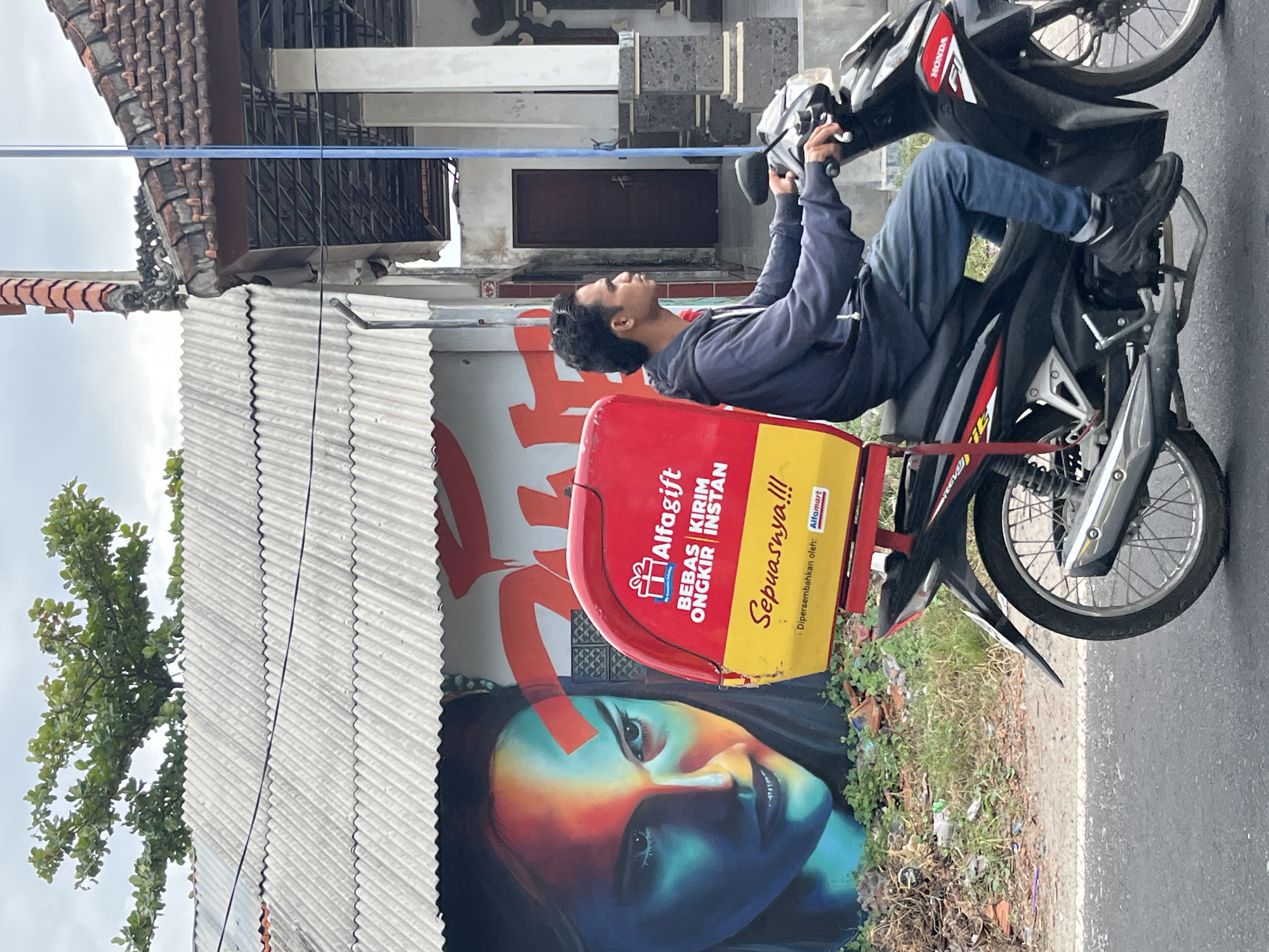 A man rides a black Honda motorcycle on a paved road in Kuta Selatan, Indonesia, with a red and yellow delivery box on the back. The delivery box has the Alfaragift logo and text in Indonesian. To the left of the road is a building with a corrugated metal roof and a large mural of a woman's face painted in vibrant colors. The mural appears to be on the side of the building. Behind the building, a tree with green foliage is visible against a cloudy sky. To the right of the motorcycle, a building with a tiled roof and a dark wooden door is partially visible. The ground to the left of the road is covered in grass and debris. The man is wearing a dark blue hoodie and blue jeans, and he is looking to the right.