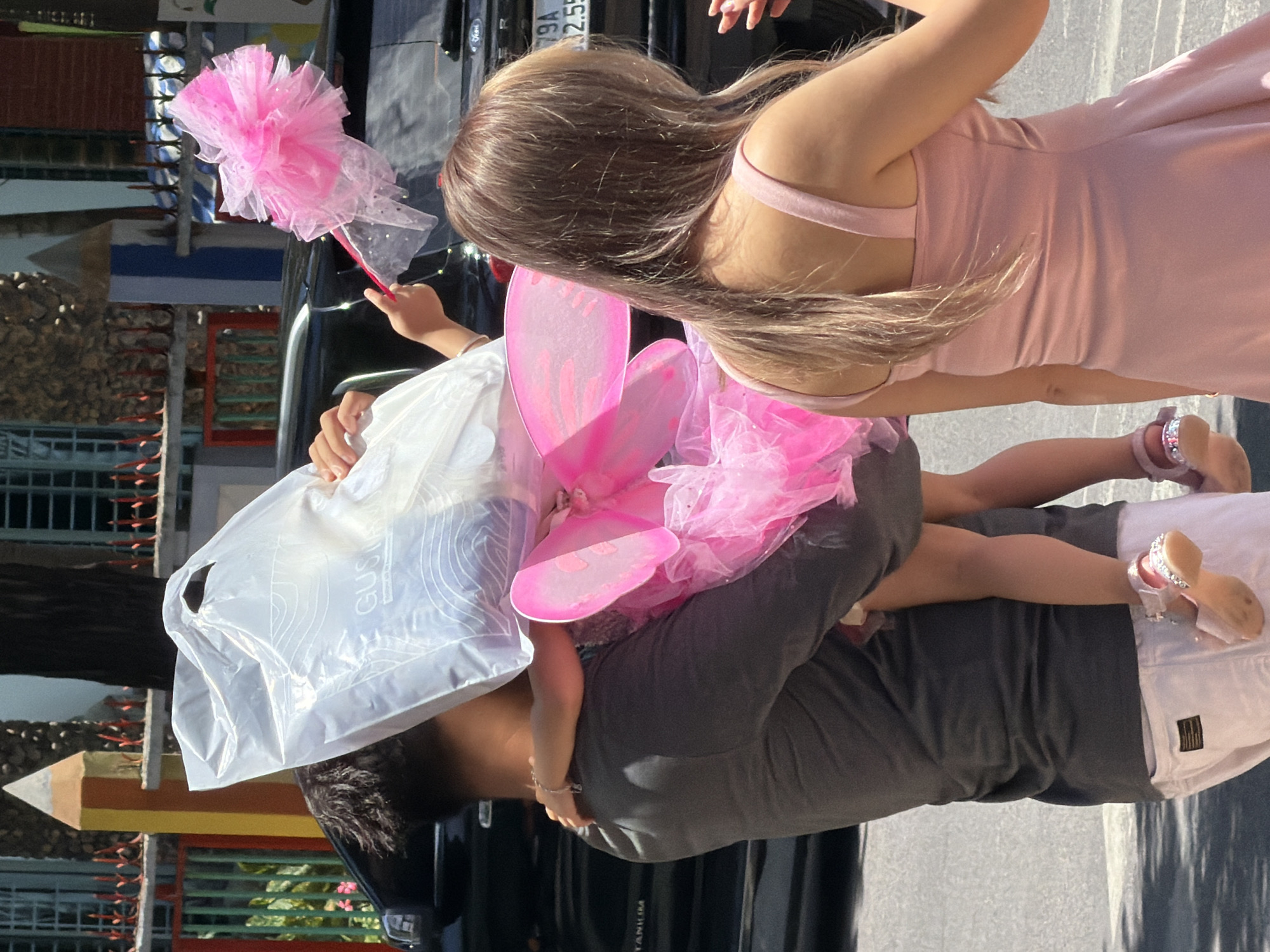 In Nha Trang, Vietnam, a man in a dark grey t-shirt and white pants carries a young child wearing a pink fairy costume with wings and a tutu. The child clutches a white plastic bag and a pink fairy wand. A woman in a light pink dress walks beside them. The scene appears to be on a street, with a black vehicle visible in the background.