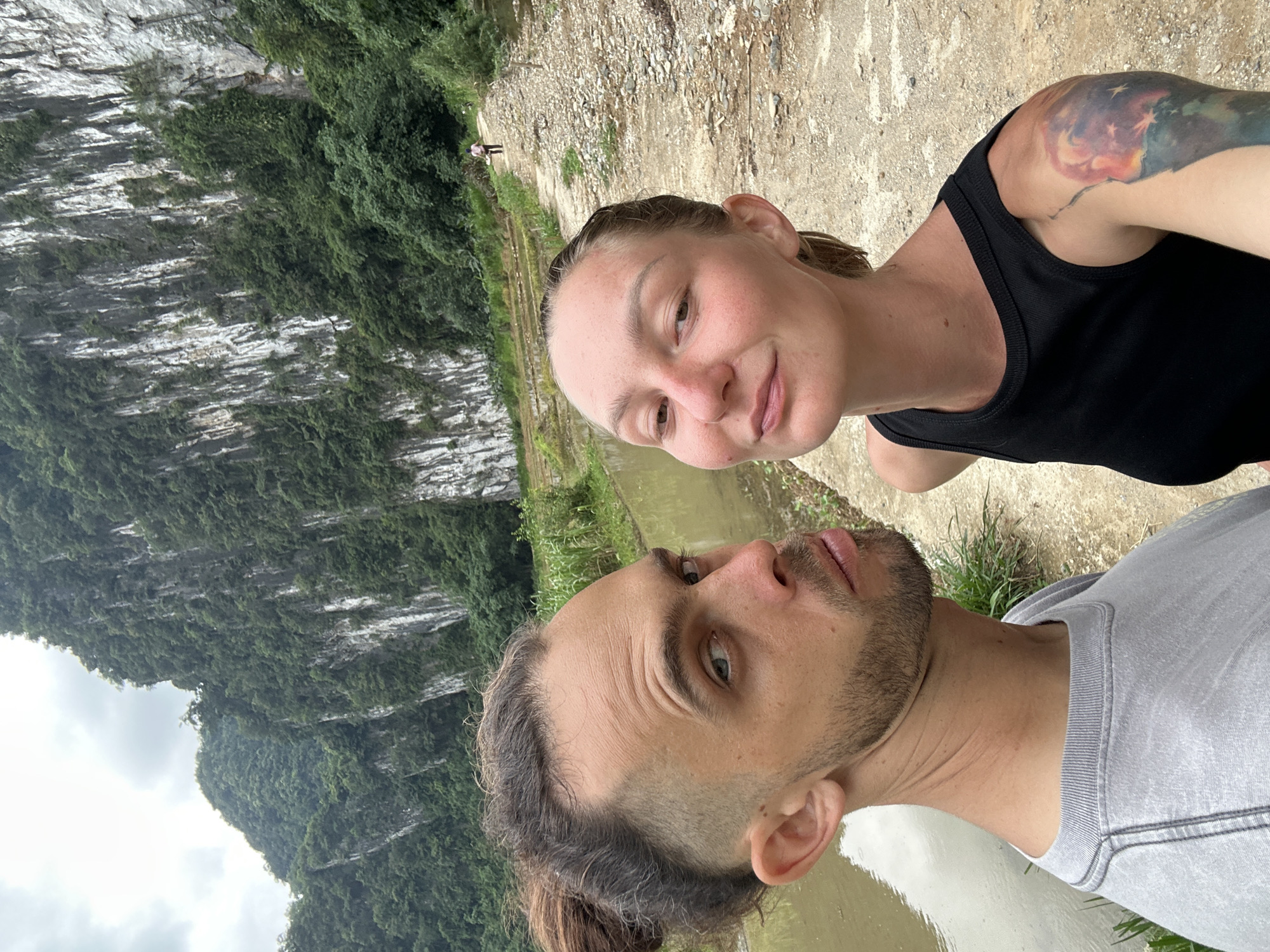 In Pù Luông Commune, Vietnam, a couple takes a selfie in a scenic natural environment. The male subject, in the foreground on the left, has his hair pulled back in a bun and is wearing a grey t-shirt. He is looking at the camera with a slightly smirking expression. To his right, the female subject is also looking towards the camera with a gentle smile. She is wearing a black tank top and has a colorful tattoo on her upper right arm.
Behind them, a dirt path stretches into the distance, flanked by a body of water on the left and lush green vegetation on the right. Towering limestone karsts covered in dense foliage dominate the background, with a cloudy sky overhead. A lone figure can be seen walking further down the path, adding a sense of depth and scale to the landscape. The scene appears to be a rural, mountainous area, likely a popular tourist destination within Pù Luông Commune.