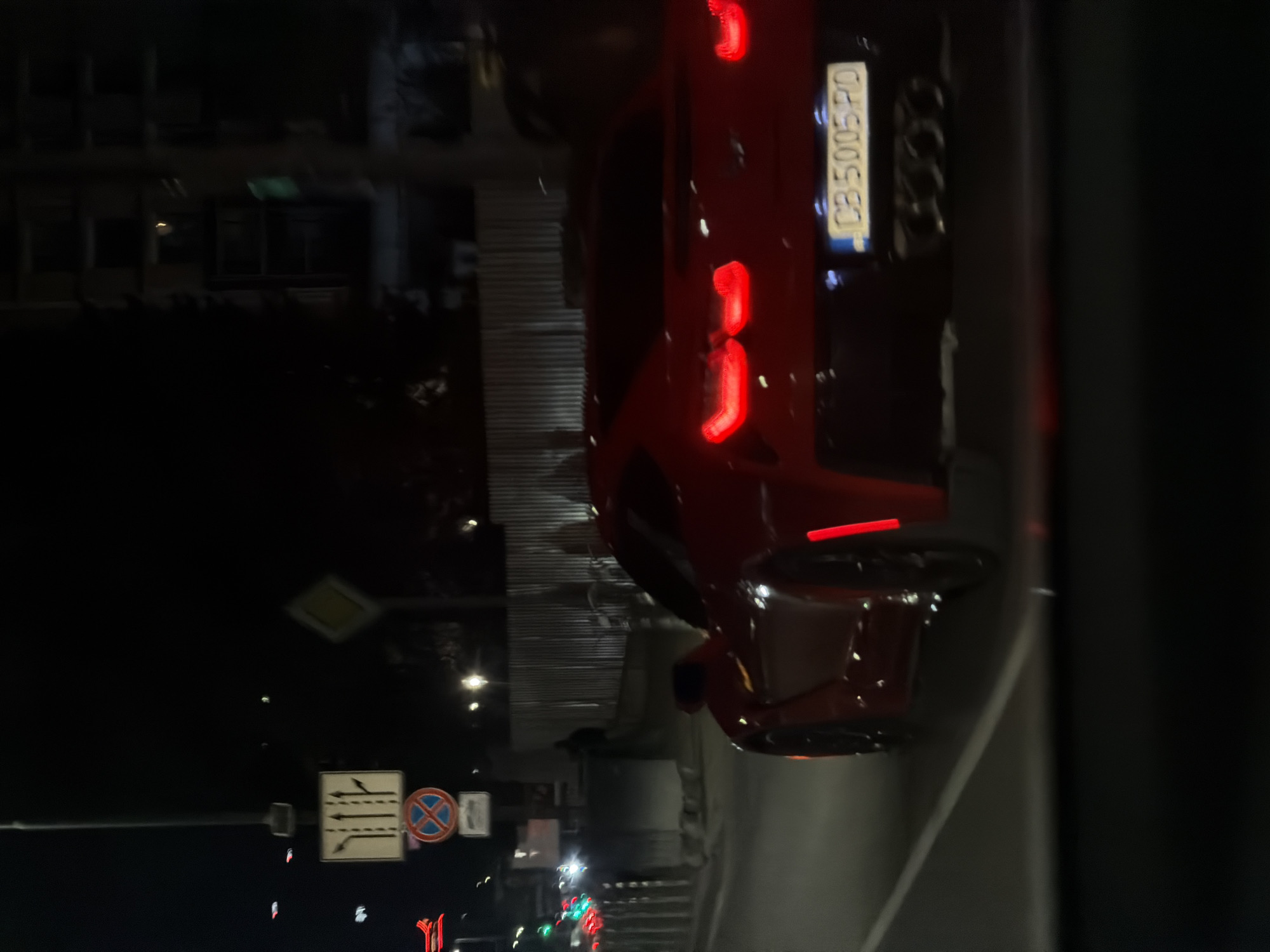 The image captures a nocturnal urban street scene in Sofia, Bulgaria, viewed from behind a vehicle. The primary subject is a vibrant red car, possibly a sports car or a sleek sedan, seen from its rear. Its LED taillights are prominently illuminated, appearing as distinct red vertical segments. The car's license plate, "CB 5005 PD," is clearly visible, with "CB" denoting the Sofia region of Bulgaria, confirming the location.
The scene is set at night, characterized by deep shadows and artificial lighting. The background features the dark silhouettes of buildings and faint glimmers from distant streetlights. Further down the road, the red and green hues of traffic lights are faintly discernible, indicating active traffic flow.
On the left side of the frame, several road signs are visible. A square white sign with black arrows indicates lane usage, suggesting directions for straight-ahead, left, and right turns. Below it, a circular "No Stopping" or "No Parking" sign (red border, blue background with a red cross) is present. Higher up, a yellow diamond-shaped sign with a white border, signifying a "Priority Road," is also identifiable.
The car appears to be either stationary or moving slowly on the street, with its bright taillights cutting through the darkness. The atmosphere is that of a typical city evening or late night. No individuals are visible in the frame. The overall impression is a factual depiction of a street at night.