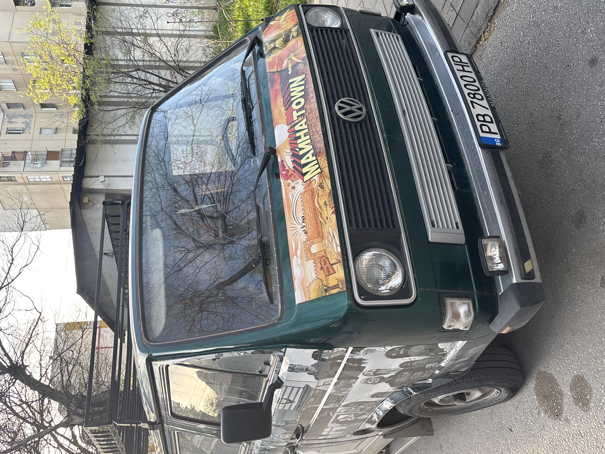 A green Volkswagen van is parked on a paved street in an urban environment. The van has a distinctive "MANHATOWN" sticker across its windshield and a Bulgarian license plate "PB 7800 HP." Reflected in the windshield are tree branches. The side of the van appears to have a black and white comic strip or patterned design. The street is paved, and there are buildings and trees in the background. The location is inferred to be in Plovdiv, Bulgaria.