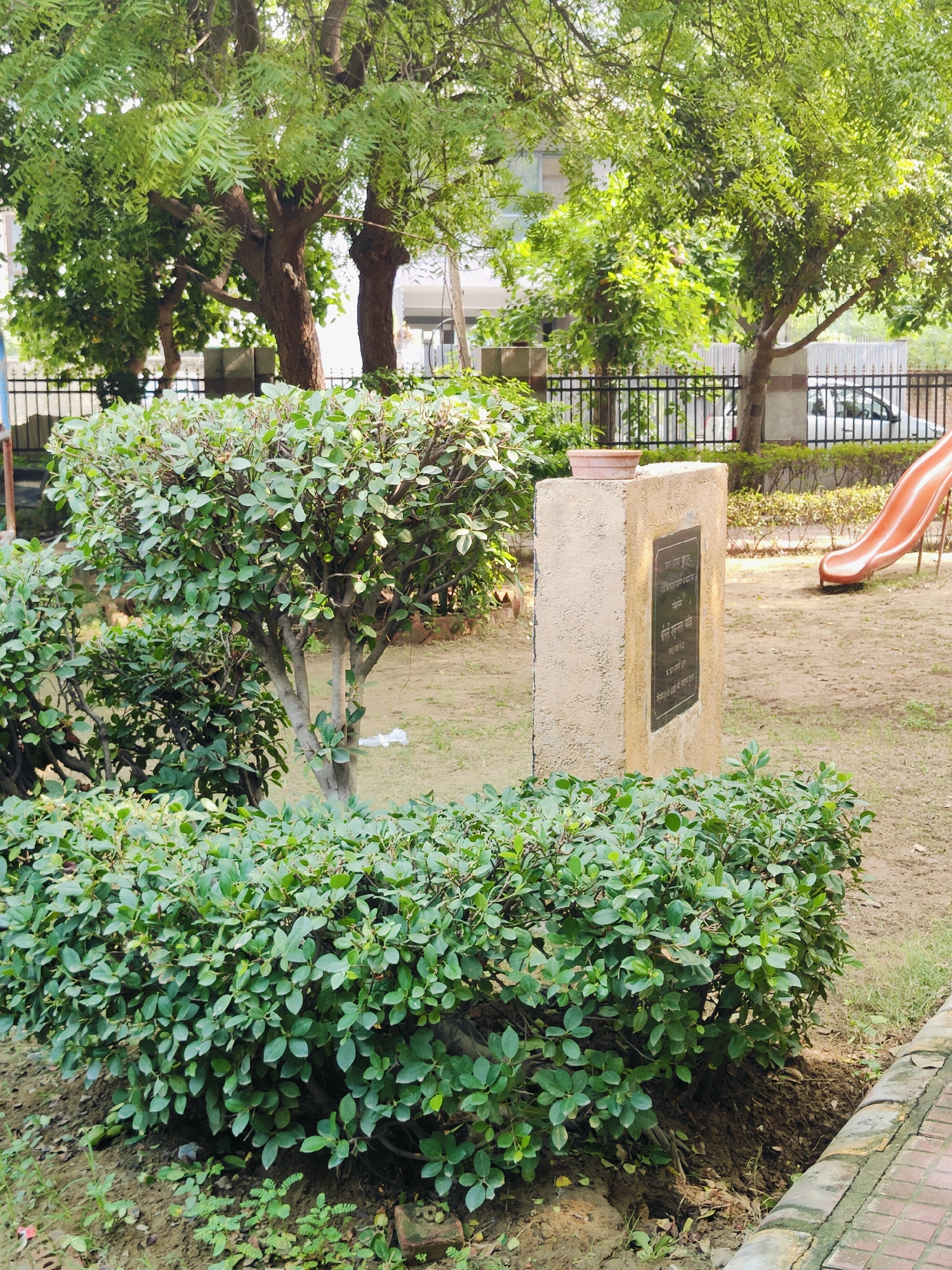 The image depicts a park area in Gurgaon, India, featuring lush green bushes and trees. In the foreground, a well-maintained, dense shrub occupies the lower portion of the frame, with a stone monument standing to its right. The monument is rectangular in shape, made of concrete, and features a dark metal plaque with engraved text, possibly a memorial or informational sign. A terracotta pot rests on top of the monument.
Behind the shrub and monument, the parkland extends, with a black metal fence visible, suggesting a boundary. Beyond the fence, trees with full green foliage provide shade. In the mid-ground, a light-colored car is partially visible, parked near a building in the background. To the far right of the image, an orange slide, characteristic of a playground, is partially visible, indicating the presence of recreational facilities. The ground cover appears to be a mix of soil and dry grass. The overall impression is that of a serene, urban green space.
