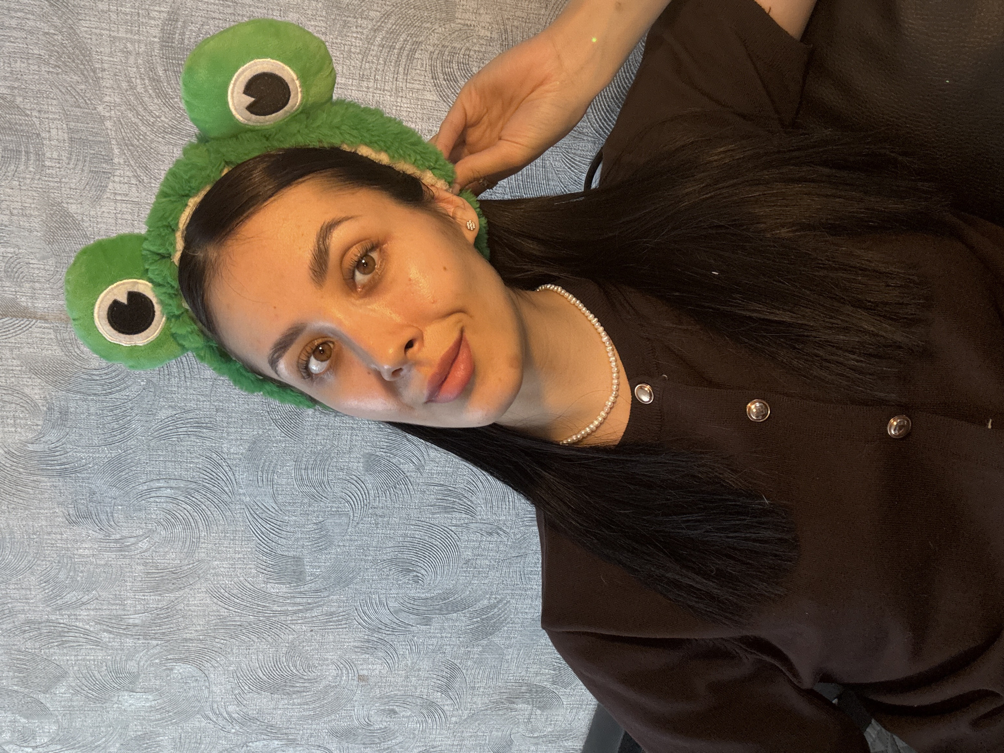 A woman is lying down on a textured, gray surface, possibly a bed or couch. She is wearing a dark brown cardigan with silver buttons and a white pearl necklace. On her head is a green headband with frog-shaped eyes. She has dark hair, light skin, and brown eyes. Her right hand is positioned near her ear. No elements indicate the location being in Votkinsk, Russia.