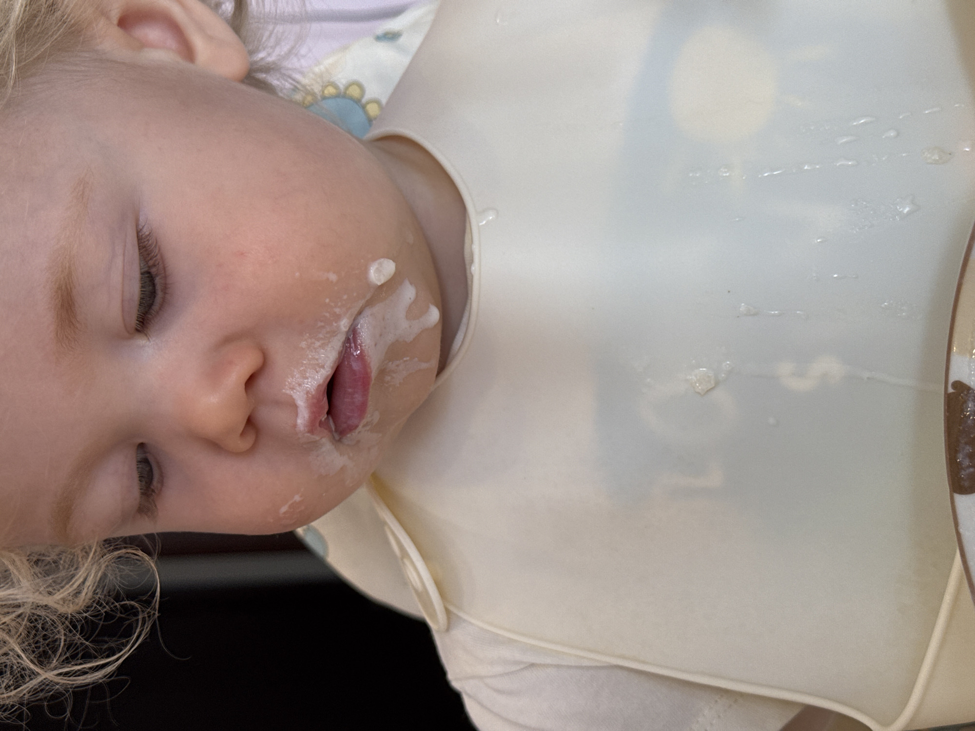A close-up shot depicts a young child wearing a white bib. The child has fair skin, blond, curly hair, and is seen with their eyes mostly closed. Residue of what appears to be food is visible on their face, particularly around the mouth. The white bib catches drips or spills. The environment is indeterminate and out of focus.