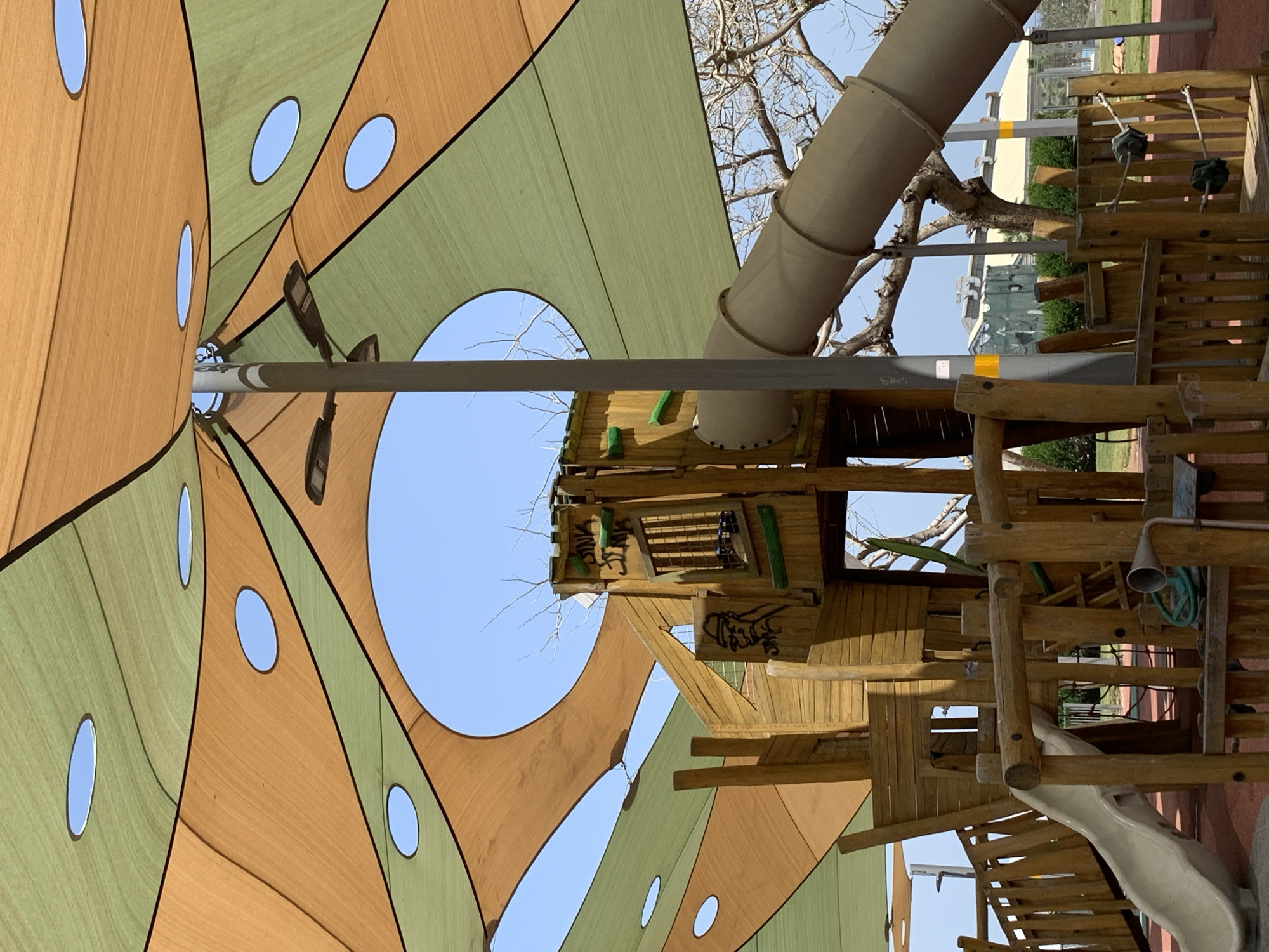 The image shows a playground structure with a large, colorful shade canopy. The canopy is green and brown with oval-shaped cutouts. A metal pole supports the canopy. Attached to the playground structure is a beige tube slide and a wooden tower with climbing features, ladders, and a slide. The background reveals buildings, trees, and a clear blue sky. The location is Tel Aviv, Israel.