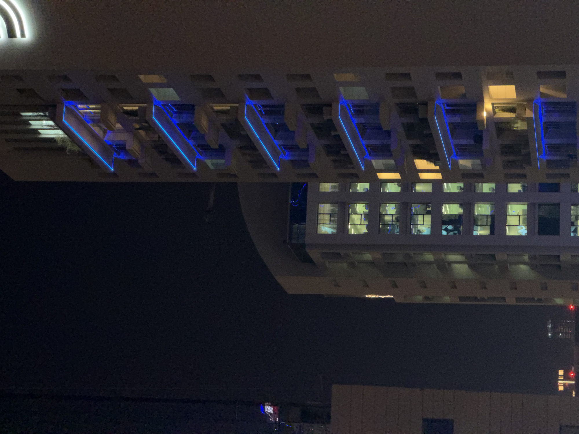 An urban nighttime scene is depicted, featuring multiple high-rise buildings illuminated by artificial light. In the foreground, positioned to the right, a tall, light-colored building stands prominently. This building features multiple cantilevered balconies on its left facade, each adorned with a bright, horizontally-oriented blue neon-like light strip running along its upper edge, creating a visually striking, layered effect down the building. Several windows on the building display internal warm yellow and white illumination. A partial, glowing white letter or symbol, possibly an 'S' or 'E', is visible at the very top right edge of this structure.
To the left and slightly behind the foreground building, a second tall, light-colored building is visible. Its windows are extensively illuminated with warm yellow-white light, indicating interior activity across many floors. A faint vertical light strip is discernible on its right side. The top of this building has a distinctive curved or rounded roof structure.
On the far left, a narrow vertical segment of a third, darker structure is present. At its upper section, a crane arm or similar construction element is identifiable, marked by small blue and red lights. Further down, a dark rectangular window or panel is visible on this structure. The sky is dark blue-black, characteristic of night, and devoid of celestial bodies. Distant, small red lights are visible at the bottom of the frame, hinting at ground-level activity. No people are visible in the image. The scene is set in an unknown location within an unspecified city.