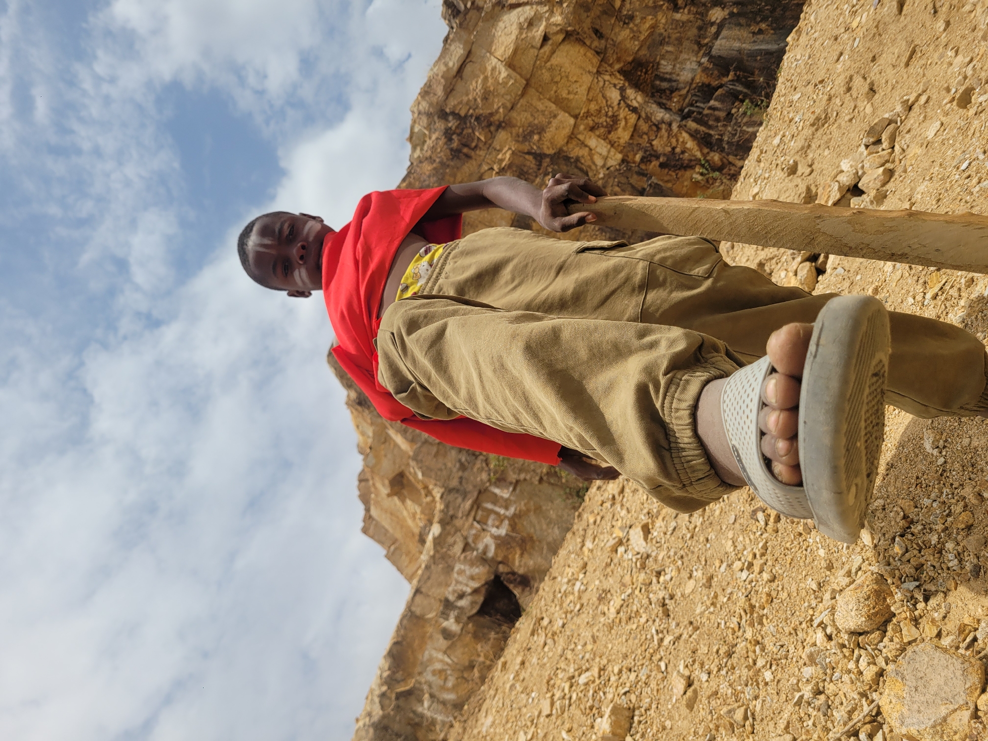 A low-angle shot depicts a young Black male standing on a rocky, sandy surface. He is wearing a red t-shirt, khaki cargo pants, and a white sandal on his right foot. His left foot is not visible. He is holding a wooden beam in his right hand. The background consists of a rocky cliff face with some vegetation. In the upper left corner, a blue sky with scattered clouds is visible. The location is Barakin Akawo, Nigeria.