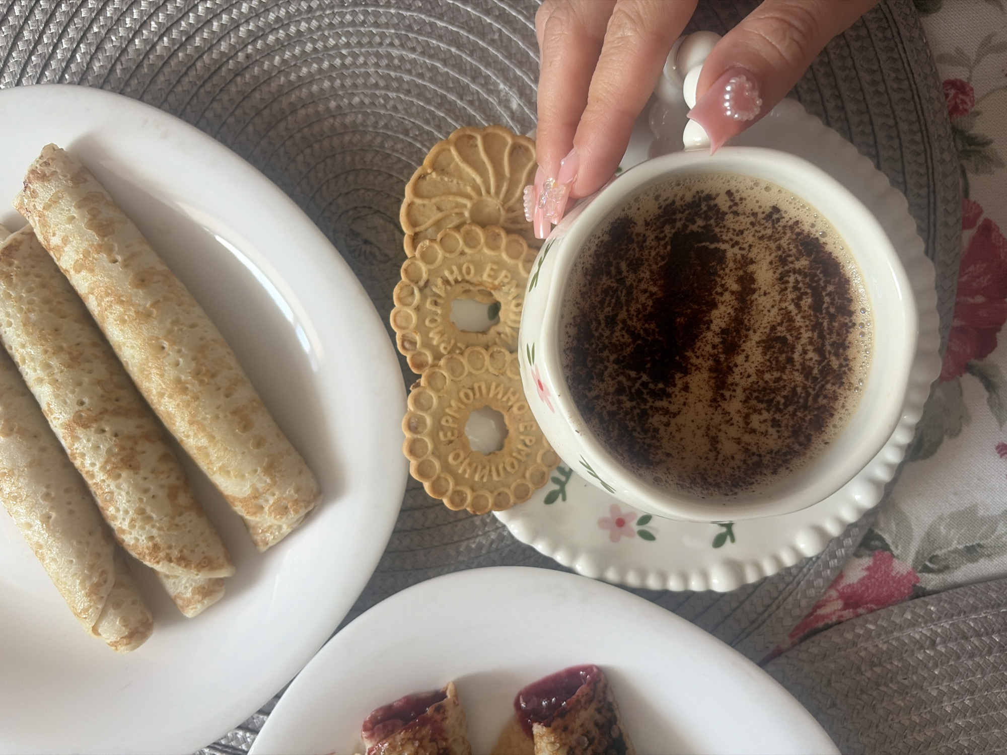 A top-down, high-angle shot captures a breakfast or snack arrangement in Saint Petersburg, Russia, featuring food and drink on a table covered by a floral tablecloth and a woven placemat. In the foreground, a white ceramic cup containing a dark beverage with a frothy top and dark powder dusting, likely coffee, is held by a female hand. The hand exhibits long, manicured nails painted in a translucent pink, adorned with small white pearls on the thumb and index finger. The cup rests on a white ceramic saucer with a scalloped edge and a subtle pink floral motif. To the right of the cup, two round, light brown cookies are visible; the prominent one has a scalloped edge and a central hole, with the brand name "ЕРМОЛИНО" (Yermolino) embossed around it. Two white plates are present: the upper plate holds three rolled items, resembling plain blini or crepes, light golden-brown with characteristic surface texture. The lower-left plate, partially cropped, contains at least two more rolled items, similar to the crepes, but with a visible dark red filling, indicative of jam or preserves, at their ends. These items are positioned on a grey, circular, tightly woven placemat, which partially covers a tablecloth featuring a pattern of pink and red roses on a lighter background.