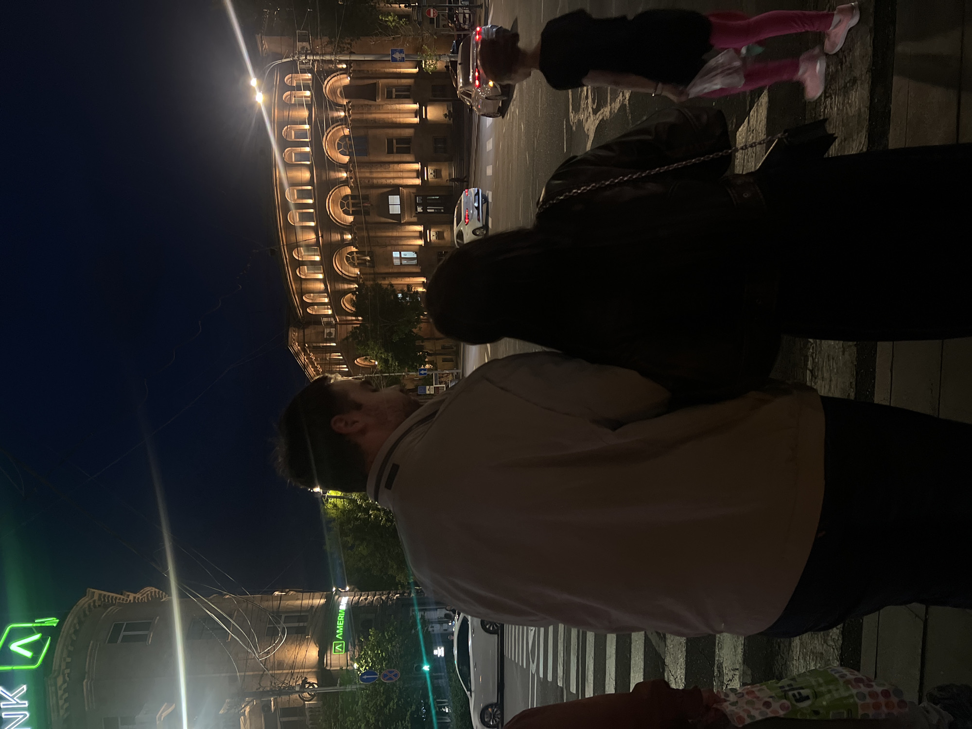 Here is a detailed description of the image:
**Scene:** Nighttime urban scene in Unknown, Unknown.
**People:**
* A man is in the foreground, seen from the back. He is wearing a light-colored jacket and dark pants.
* A woman is partially visible, standing next to the man, wearing a dark coat.
* A child is seen running in the background, wearing a dark top and bright pink leggings.
* A baby is in a stroller near the bottom of the image.
**Objects:**
* A large building with illuminated arches is visible in the background.
* An "Ameriabank" sign is seen on a building to the left.
* Cars are moving along the street.
* A stroller
**Actions/Interactions:**
* The man appears to be looking at something in the distance, possibly the illuminated building.
* The child is running across the street.
* The group is standing on a crosswalk.
**Environment:**
* It is nighttime, and the scene is illuminated by streetlights and the building's lighting.
* Buildings line the street.
**Location:** The scene appears to be a street corner in Unknown, Unknown, with a prominent building in the background and a bank visible on the left. A crosswalk is on the road.