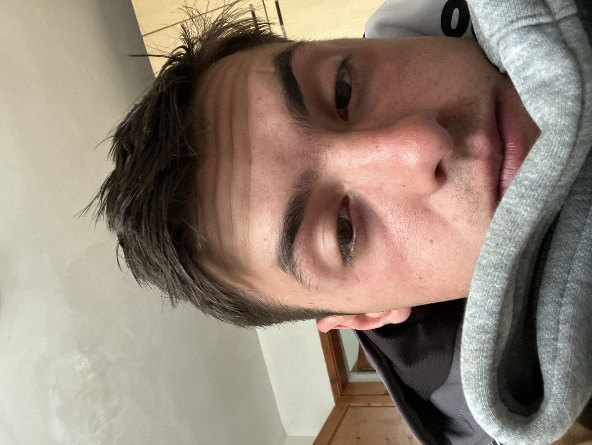 A close-up, slightly rotated self-portrait of a Caucasian male. He has dark brown hair, light skin, and dark eyes. His hair is short and slightly messy. He is wearing a gray hooded sweatshirt with the hood partially obscuring his face. The background is blurry and indistinct, showing a beige-colored wall and a wooden frame or door. There is no indication that the photograph was taken in the city of Krastevich, Bulgaria.