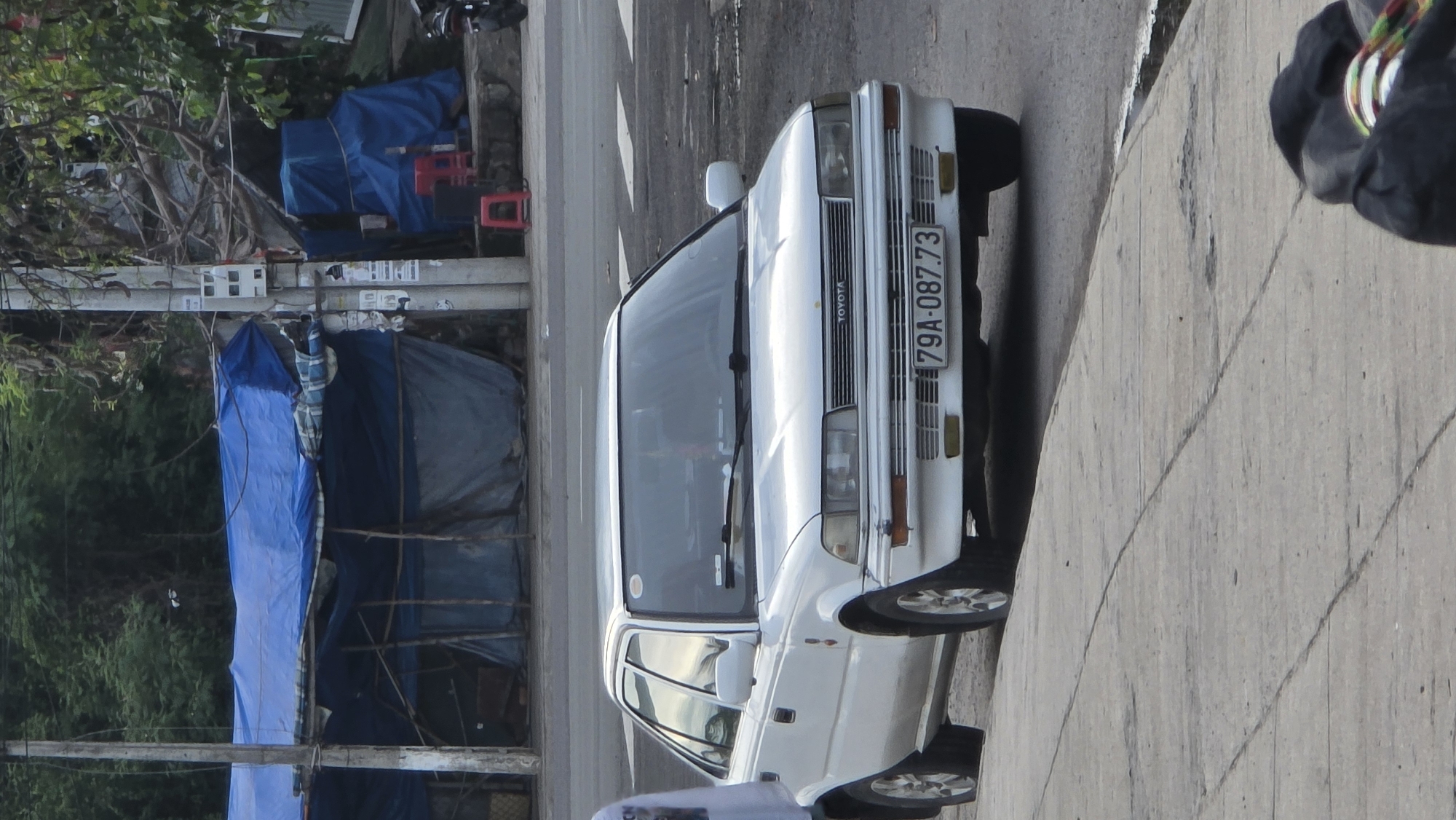 A white Toyota sedan is parked on the right side of a paved road in Bac Nha Trang Ward, Vietnam. The car, an older model, features a visible "TOYOTA" logo on its front grille and a Vietnamese license plate displaying "79A-087.73". Its headlights are rectangular, and its body is clean white.
To the left of the car, the background is characterized by a mix of greenery and human-made structures. Several utility poles, covered with various papers and posters, stand tall. Behind these, numerous structures, possibly temporary shelters or storage areas, are covered with blue tarpaulins. Green trees and dense foliage are visible above and behind these structures. A few red plastic stools are also discernible among the background elements.
On the far right edge of the frame, a person's lower leg and foot, clad in dark trousers and shoes with colorful laces, are visible on what appears to be a concrete sidewalk or shoulder. The road surface in the foreground shows some wear and cracks. The lighting suggests it is daytime, possibly with diffused sunlight. The scene depicts a typical street environment, combining urban infrastructure with elements of informal construction.