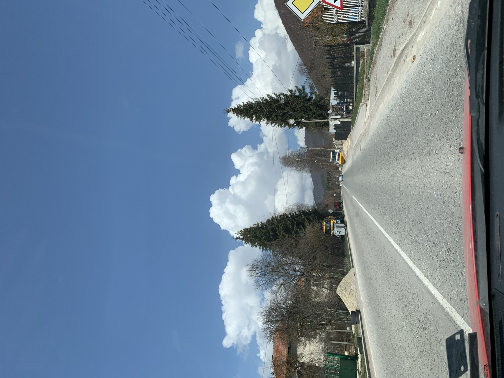 The image captures a rural road scene in Gintsi, Bulgaria, viewed from inside a moving vehicle. The asphalt road stretches into the distance, marked by a prominent solid white line on its right side. On the left of the road is a dirt or gravel shoulder, beyond which are various trees and buildings.
The sky is a clear, vibrant blue, adorned with large, fluffy white cumulus clouds. Multiple power lines traverse the upper portion of the sky. Along the roadside, there's a mix of vegetation, including tall evergreen trees with full, dark green foliage and several deciduous trees that are currently bare, suggesting the season is late winter or early spring. Behind the trees, several houses with characteristic reddish-brown tiled roofs and fences are partially visible.
Further down the road on the left, a yellow construction vehicle, appearing to be an excavator or similar heavy equipment, is parked or positioned near the roadside, accompanied by a smaller white utility vehicle. In the upper left quadrant of the image, a portion of a yellow diamond-shaped road sign with a black border, typically indicating a priority road, is visible. Another road sign is partially obscured next to it. The entire scene is bathed in bright daylight, indicating a sunny day. The right edge of the frame shows elements of the vehicle from which the photograph was taken, including a red painted surface and a black panel with some small, illegible white marks. No explicit text is clearly discernible on any signs or objects.